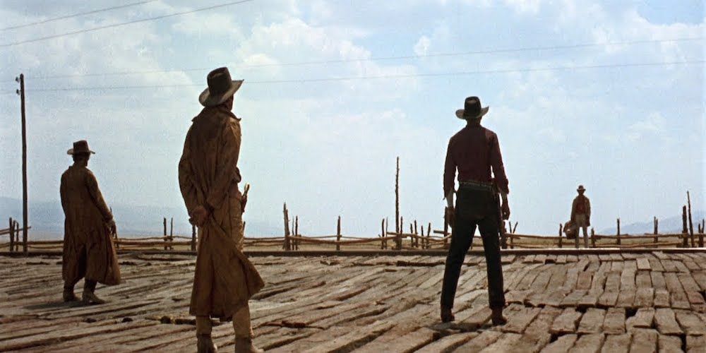 3 men wearing long dusters stare at another man holding a bag across the train tracks