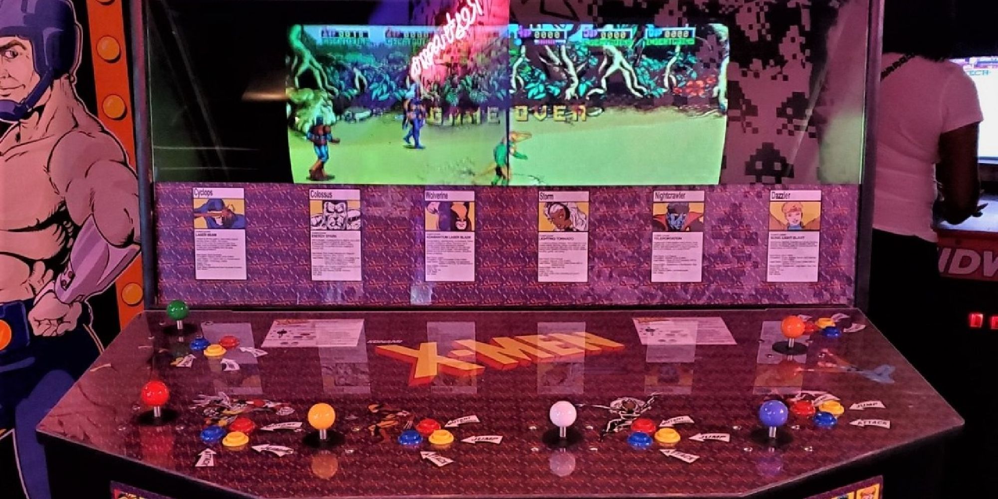 A photo of the 1992 X-Men arcade cabinet, showing its six player inputs and the mirrored double-wide screen