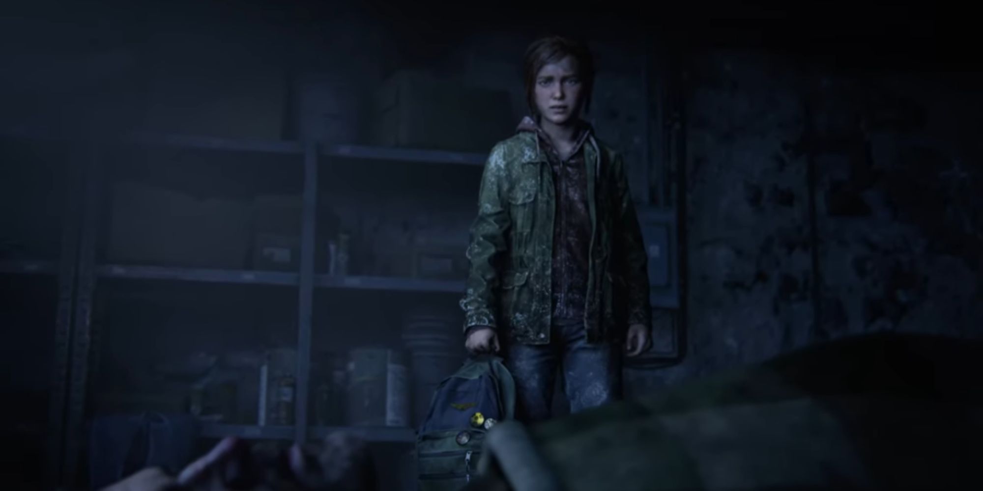 Ellie stands over Joel in a dark garage with a backpack in her hands
