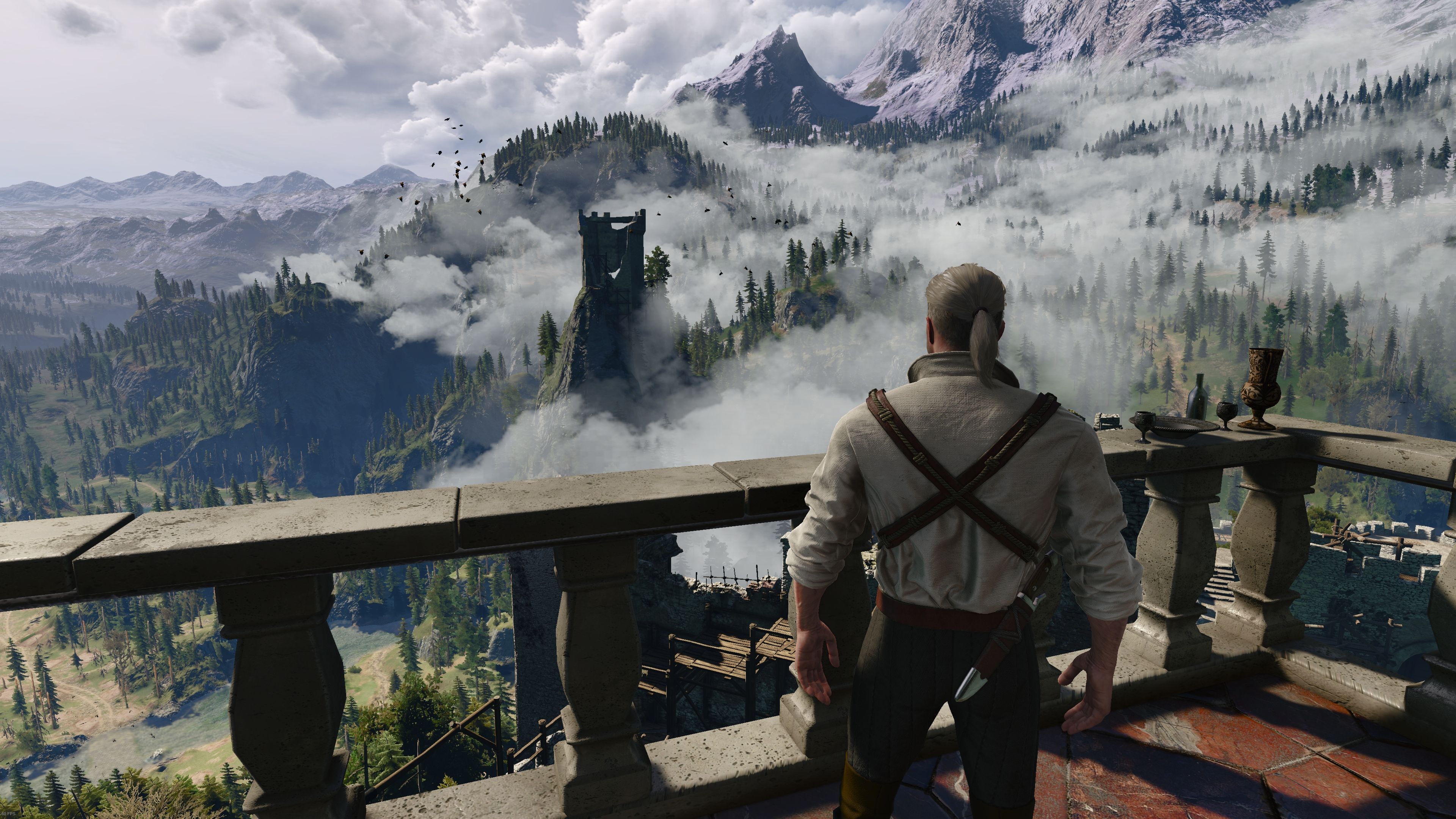 A man dressed in a white linen shirt looks out of a balcony and over a mountain pass shrouded in clouds.