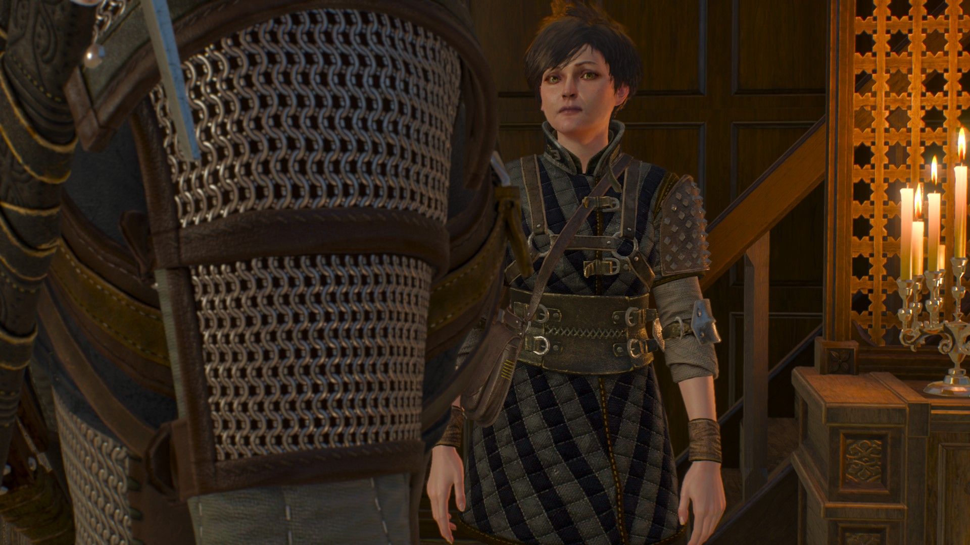 A young woman with short hair wearing witch hunter armor talks to Geralt inside an Oxenfurt home.