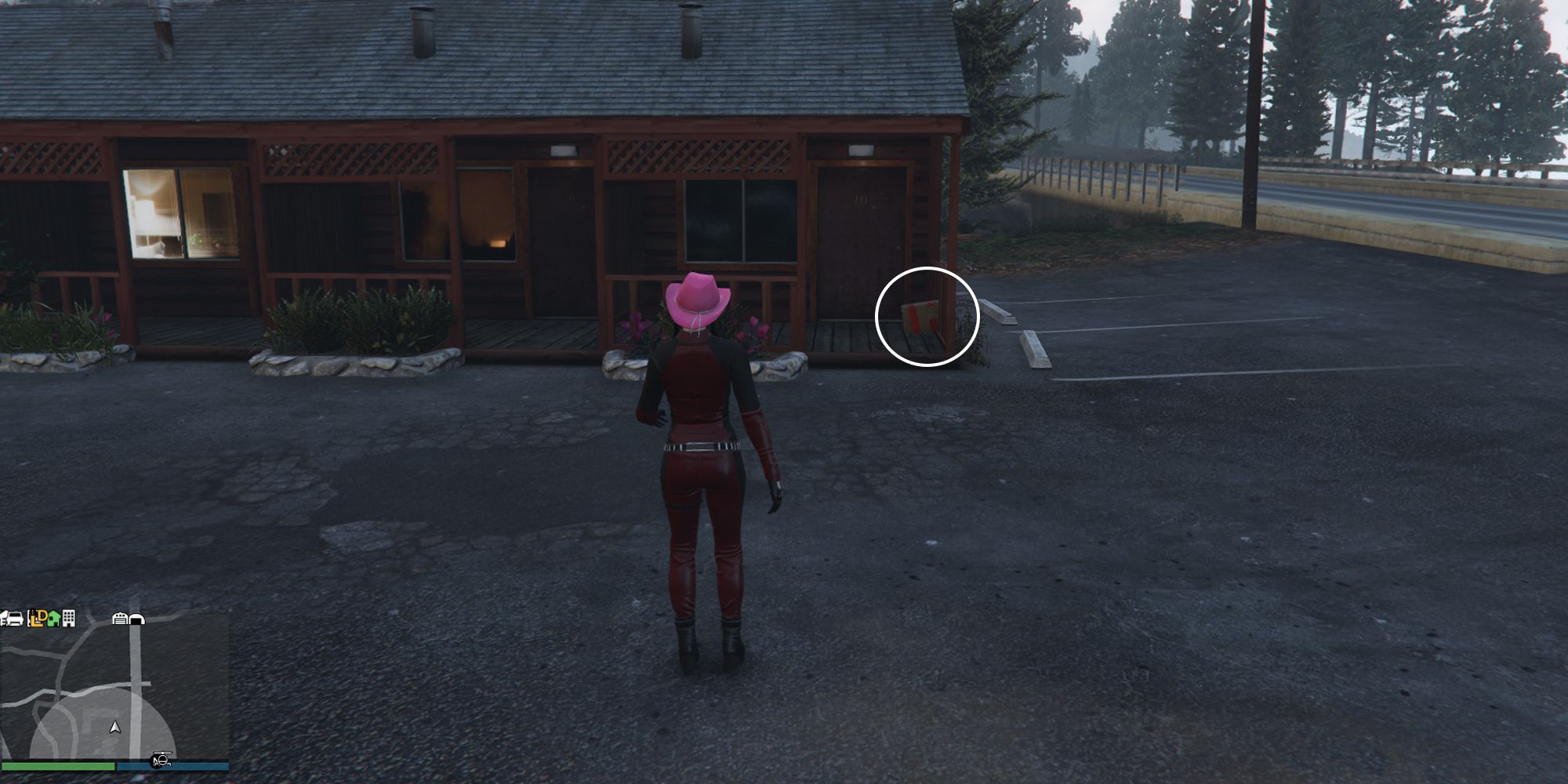 Image depicts a female character in GTA Online at Bayview Lodge looking towards the row of lodges next to the main road. In the entrance of the lodge that is the furthest to the right, the box of G's Cache is sat on the decking.