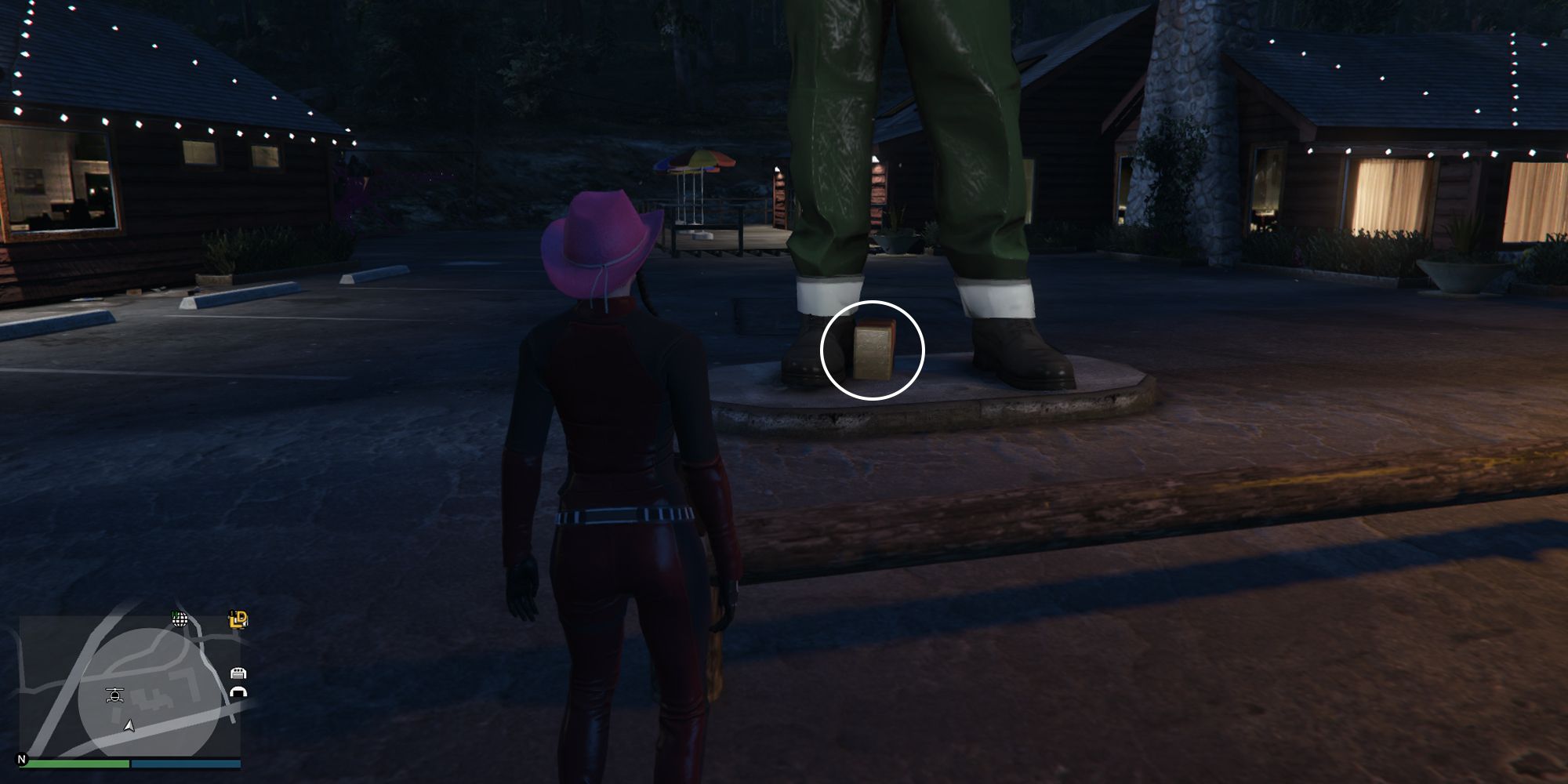 Image depicts a female character in GTA Online at Bayview Lodge stood in front of the feet of a large lumberjack statue. In between the statues feet is the G's Cache box.