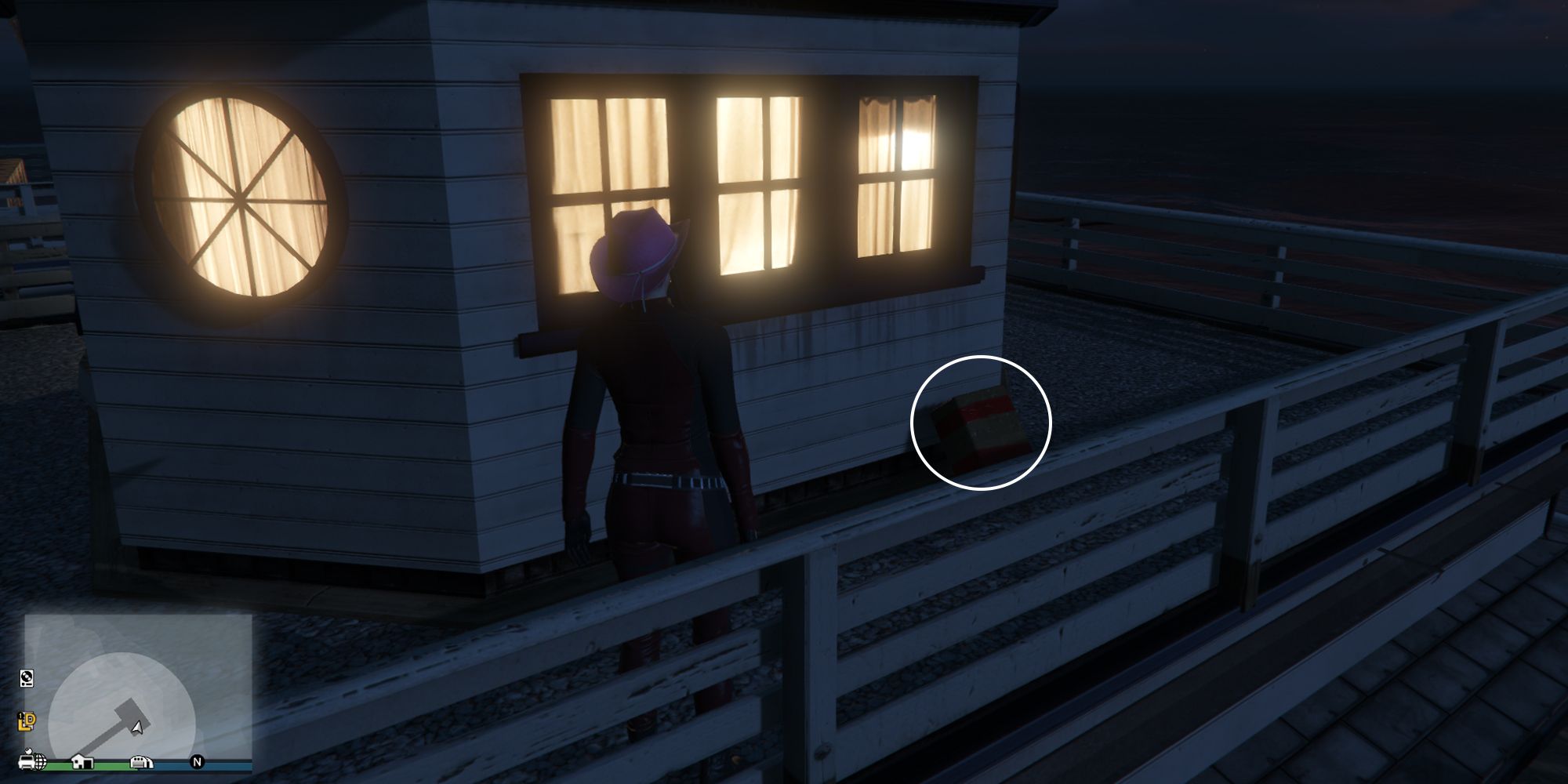 Image depicts a female player in GTA Online at the Chumash Historic Family Pier on the upper level of the buildings at the end of the dock. The lights are on in the house, and G's Cache is on the floor resting next to the building.