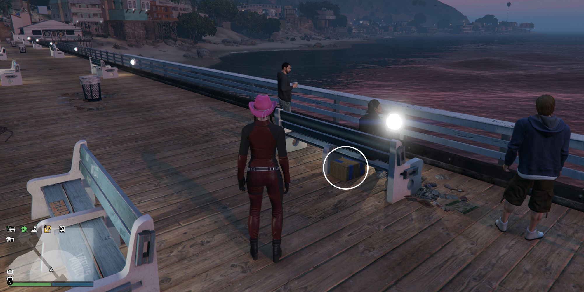 Image depicts a female player in GTA Online at the Chumash Historic Family Pier stood next to a bench with three male NPC's stood near to it. There are rows of benches going down the pier behind this bench. Underneath this bench is G's Cache.