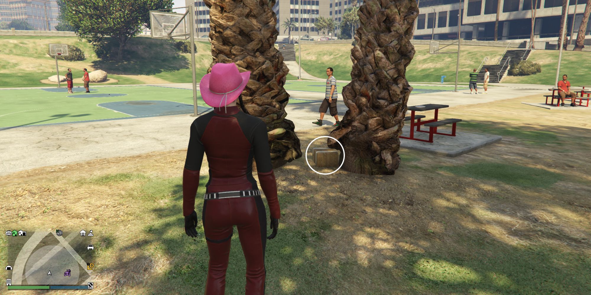 Image depicts a female player in GTA Online at Decker Park stood facing two trees next to a series of red benches. There is a small gap between these two trees, and G's Cache is on the ground in between them. There are also NPC's in the park in the background.