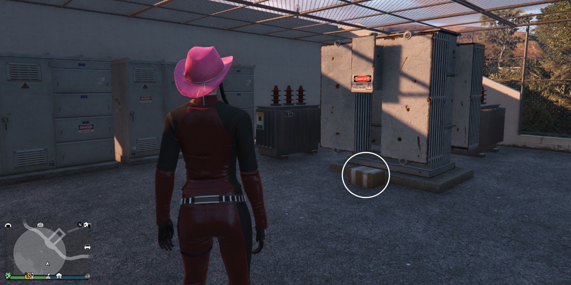 Image depicts a female player in GTA Online at Galileo Observatory in a fenced off maintenance area surrounded by trees. There are power boxes and generators in this area. G's Cache is on the floor in front of one of the big power boxes. 