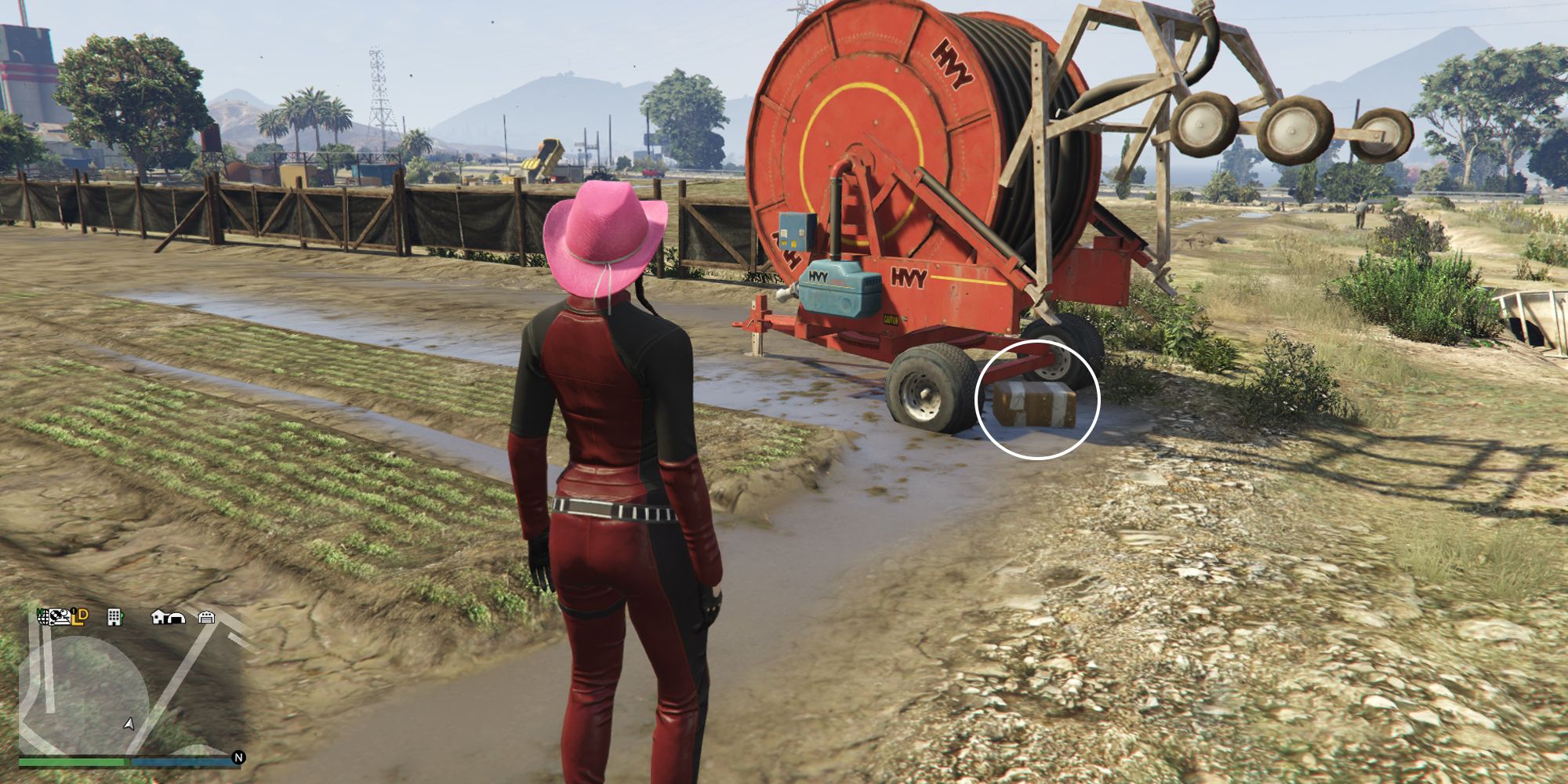 Image depicts a female character in GTA Online at Grapeseed Rail Terminus stood in front of a piece of red farming equipment in a field. Underneath the front tire on the equipment is G's Cache box.