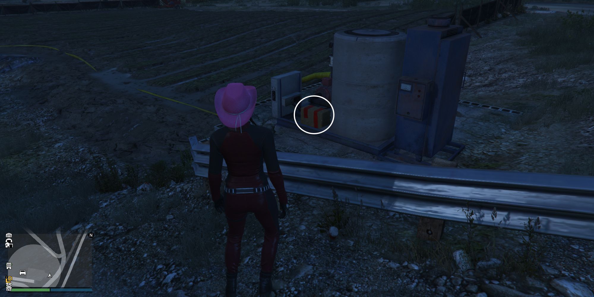 Image depicts a female character in GTA Online at Grapeseed Rail Terminus stood next to a water supply system overlooking some farming fields. Next to some of the water pipes is G's Cache.