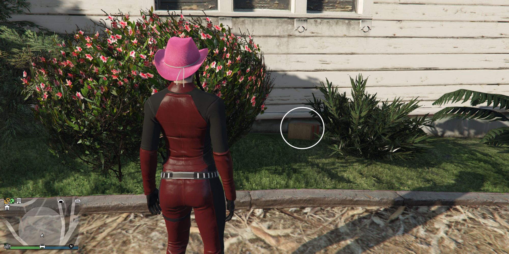 Image depicts a female character in GTA Online at Hill Valley Church stood in front of one of the side walls of the building, with some flowers and bushes around. In between a big bush of flowers, and a smaller green bush is G's Cache.