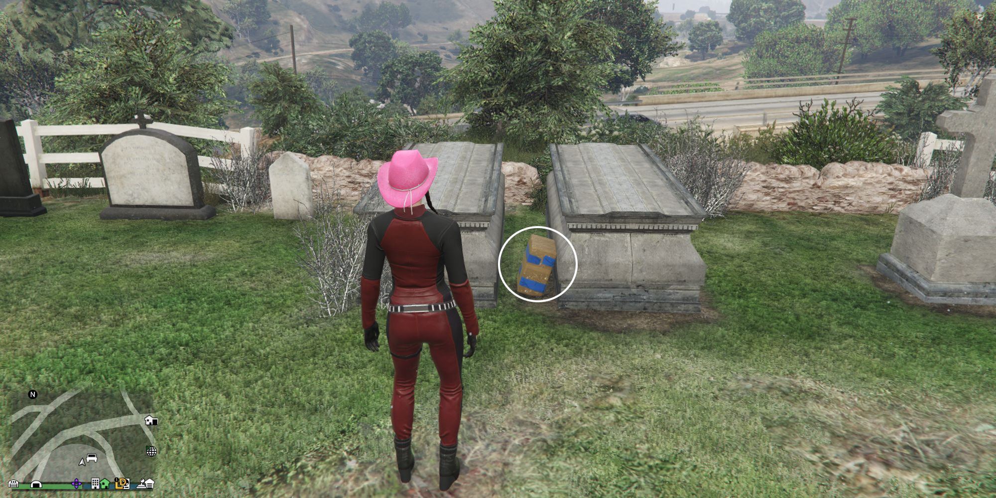 Image depicts a female character in GTA Online at Hill Valley Church stood in front of two tombs in the church graveyard. There are other tombstones and memorials around. In between the two large tombs is G's Cache.