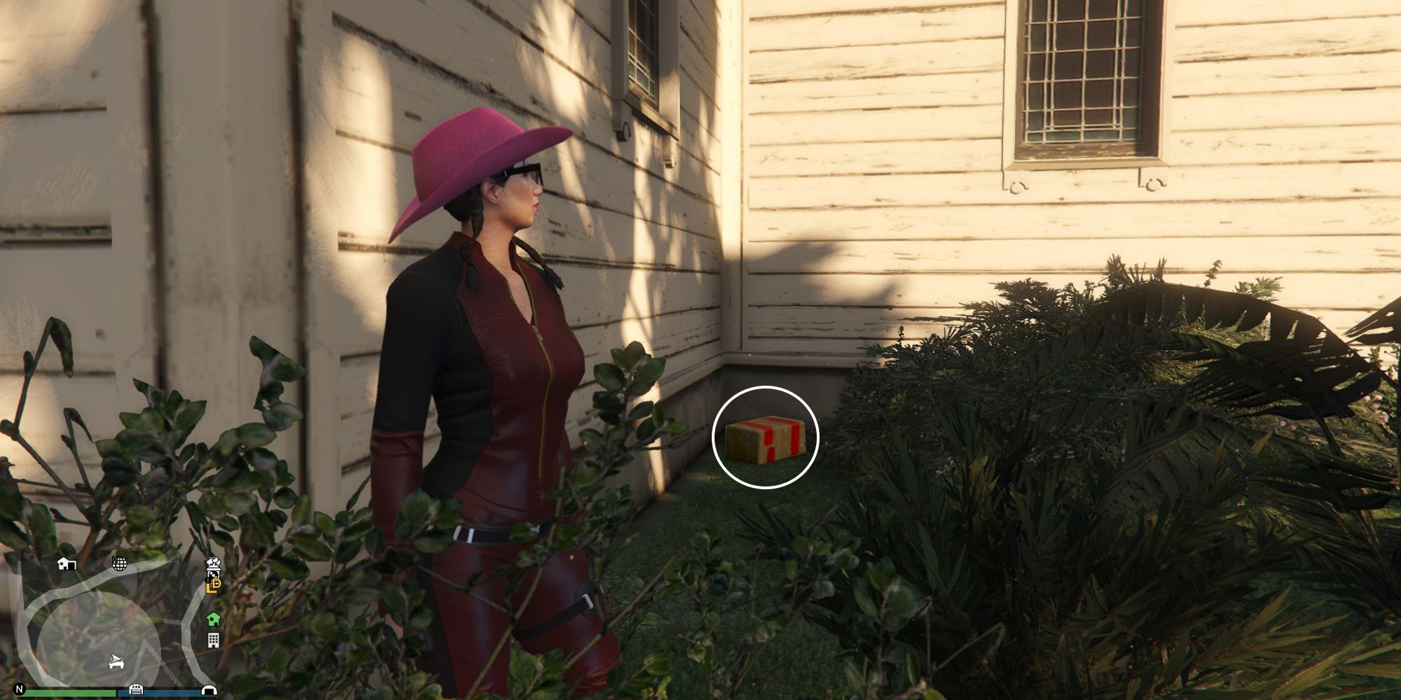 Image depicts a female character in GTA Online at Hill Valley Church facing away from the main church building among some bushes and flowers. This side of the building has two windows, and G's Cache is on the floor underneath the leftmost window.