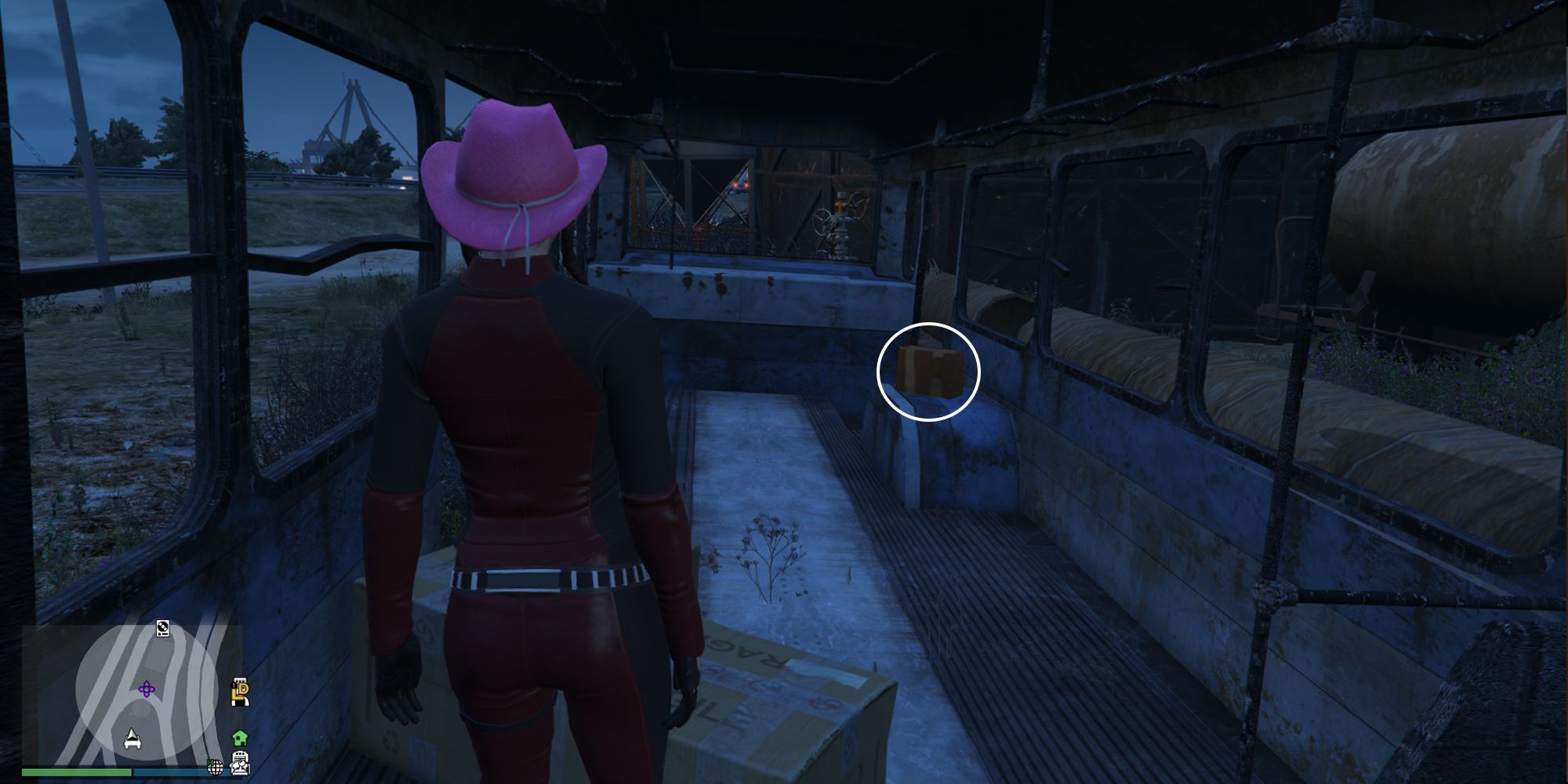 Image depicts a female player in GTA Online at Jay and Sons in El Burro Heights stood inside an old abandoned bus. In the bus are a few boxes, and some small spaces where seats have been removed. G's Cache is on a small raised platform inside the bus.