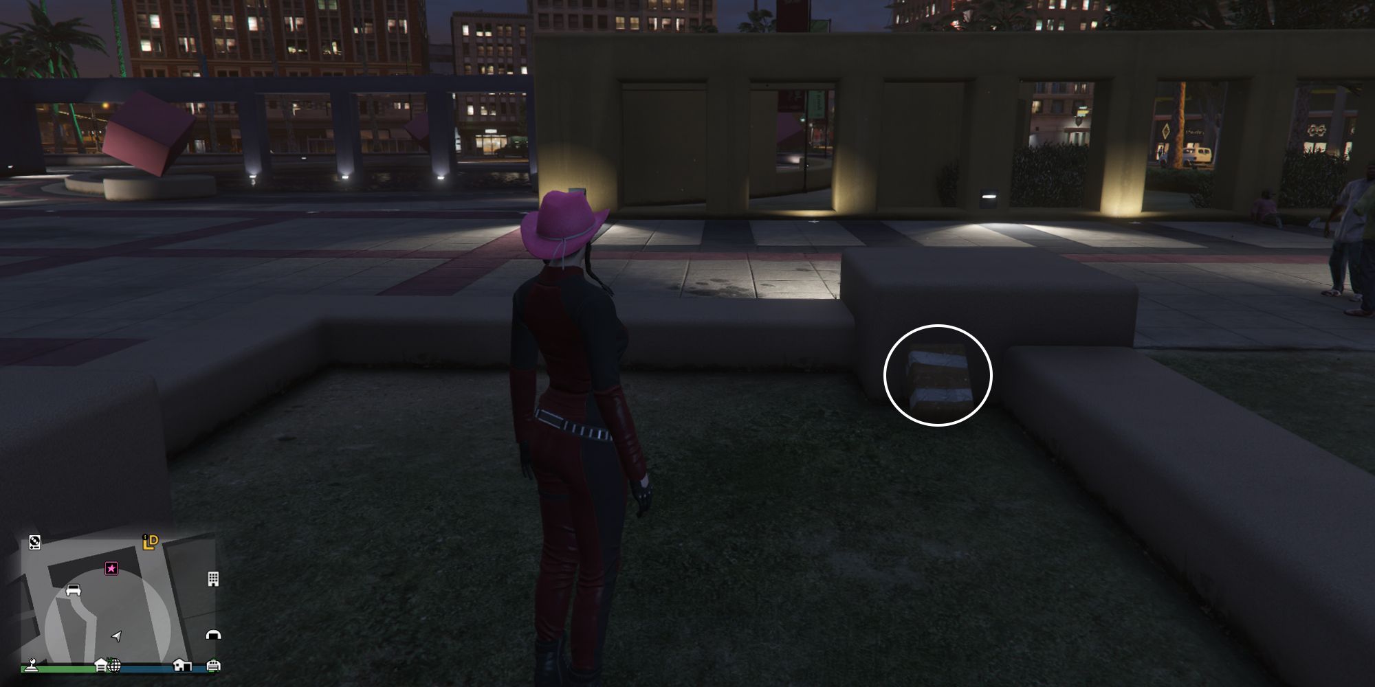Image depicts a female player in GTA Online at Legion Square stood in a small patch of grass surrounded by a short grey wall. In the background, there are larger yellow walls with gaps in it, and a fountain with a large red cube in the middle. G's Cache is to the right of the player, propped against the grey wall.