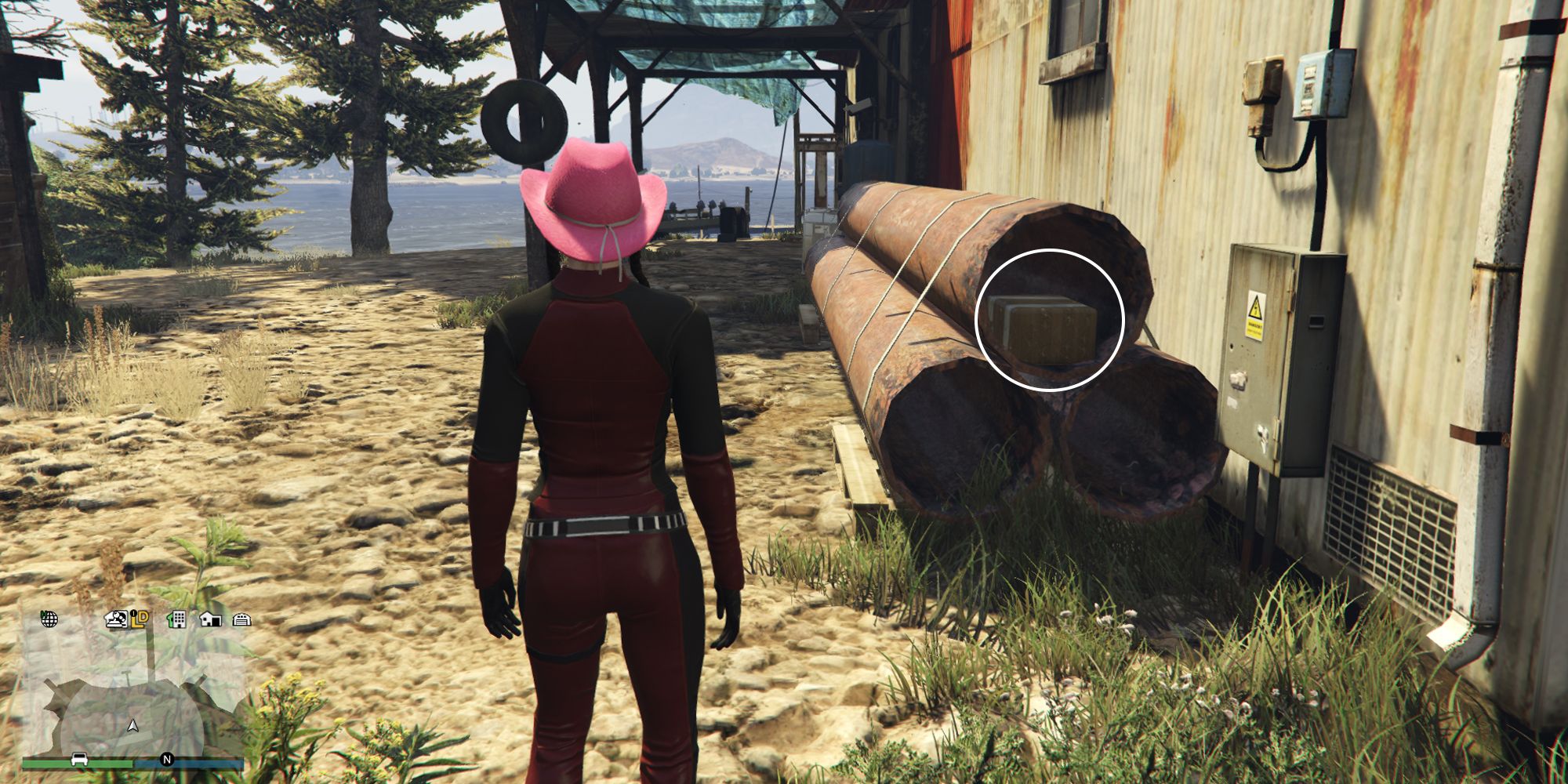 Image depicts a female character in GTA Online at Millar's Fishery stood in front of another building that overlooks the water. To the right, there are three metal pipes stacked on top of each other, and G's Cache is in the pipe on the top of the pile. 