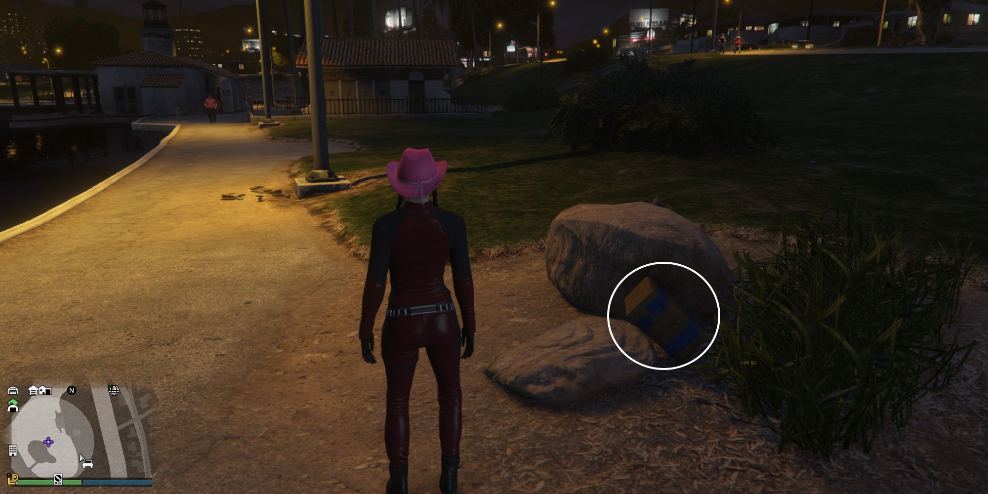 Image depicts a female player in GTA Online at Mirror Park Lake with the lake on the left, a toilet block in front of them, and two rocks on the right. In between these two rocks, G's Cache is sat on them.