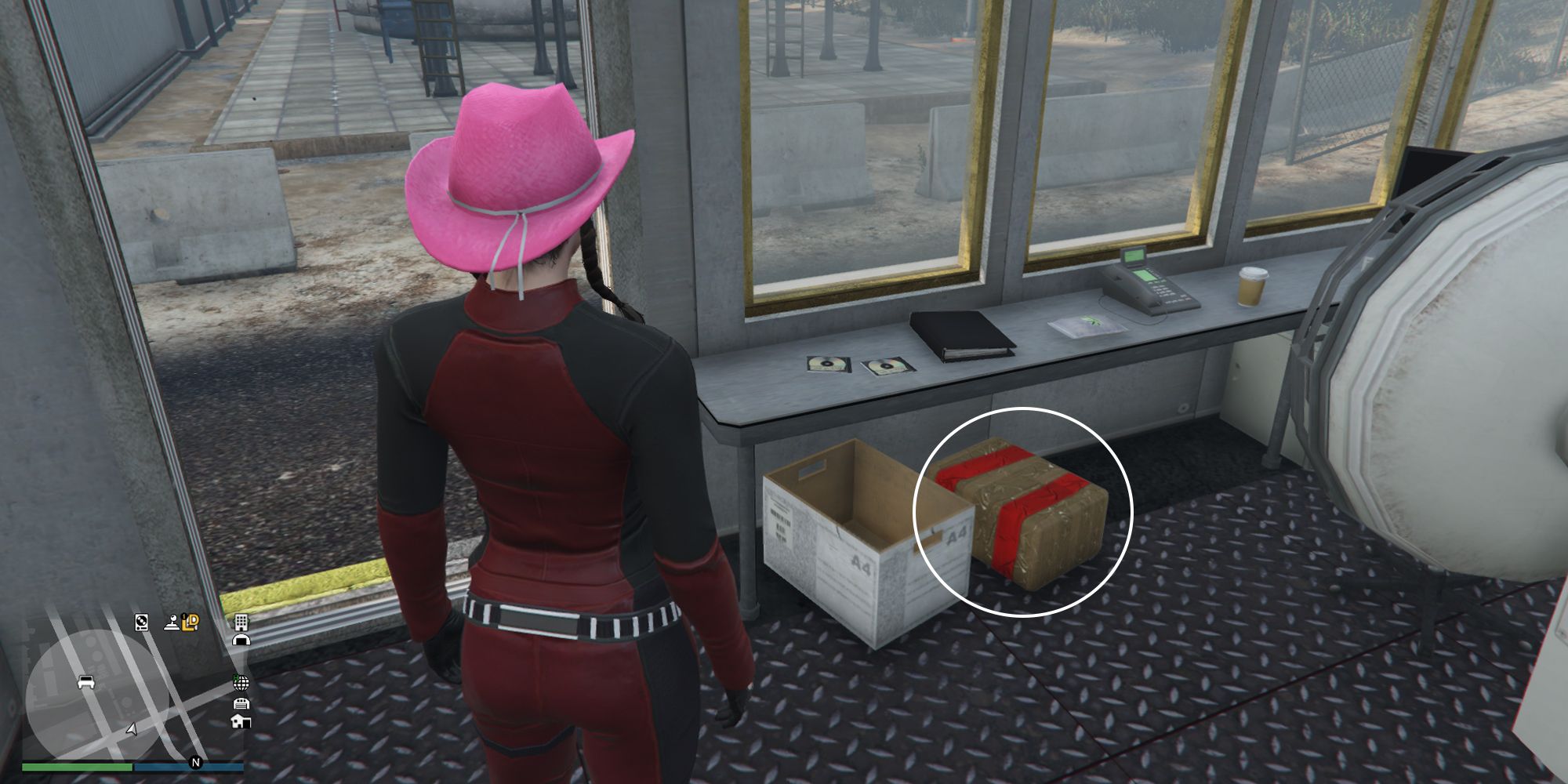 Image depicts a female character in GTA Online at Palmer-Taylor Power Station inside the security checkpoint room. Inside are some office supplies on a desk, a white box underneath the desk with G's Cache box next to this.