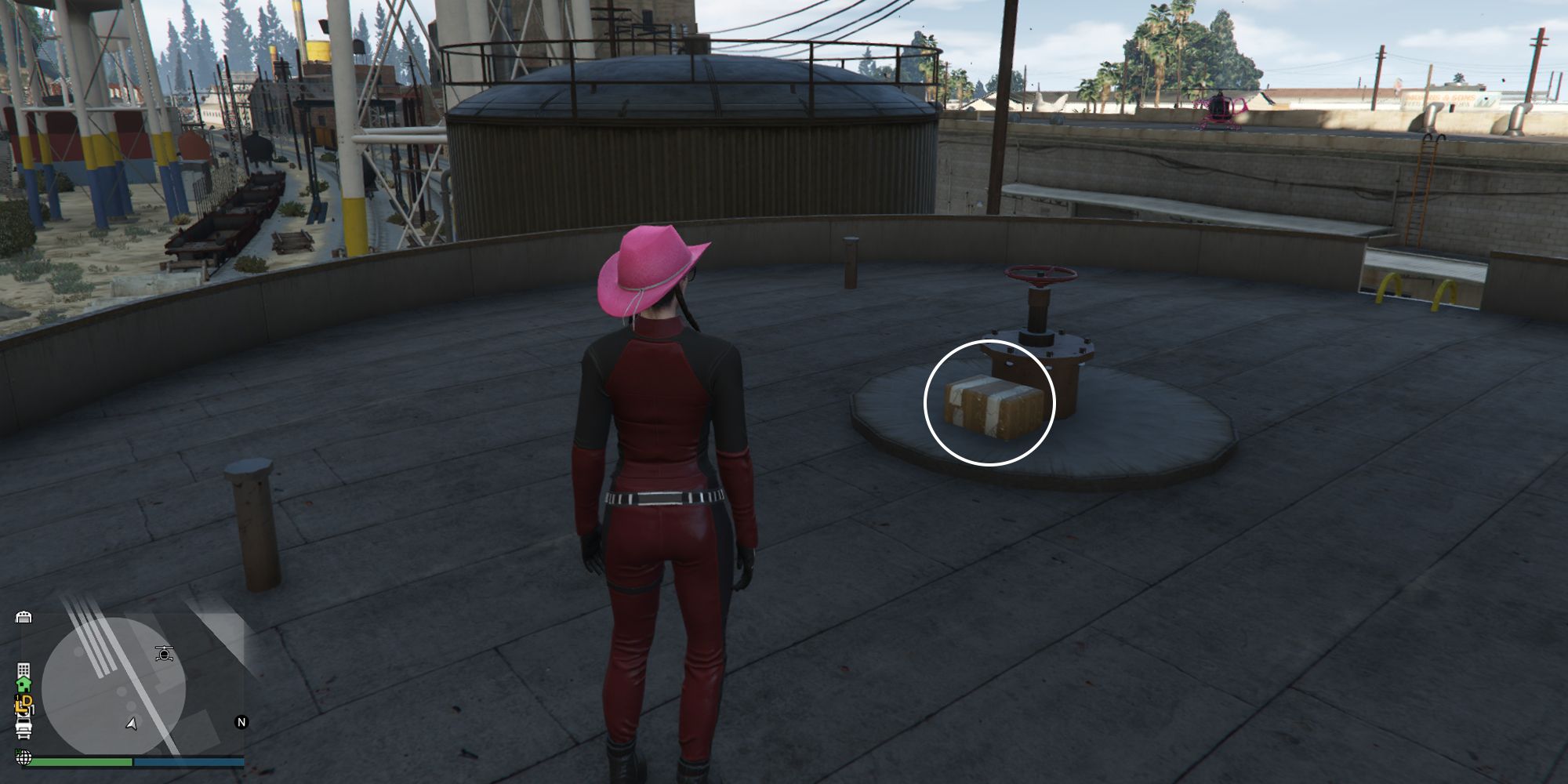 Image depicts a female character in GTA Online at Railyard Clucking Farms on top of a large metal tank overlooking the rest of the railyard. In the centre, the G's Cache box is sat against a pipe.