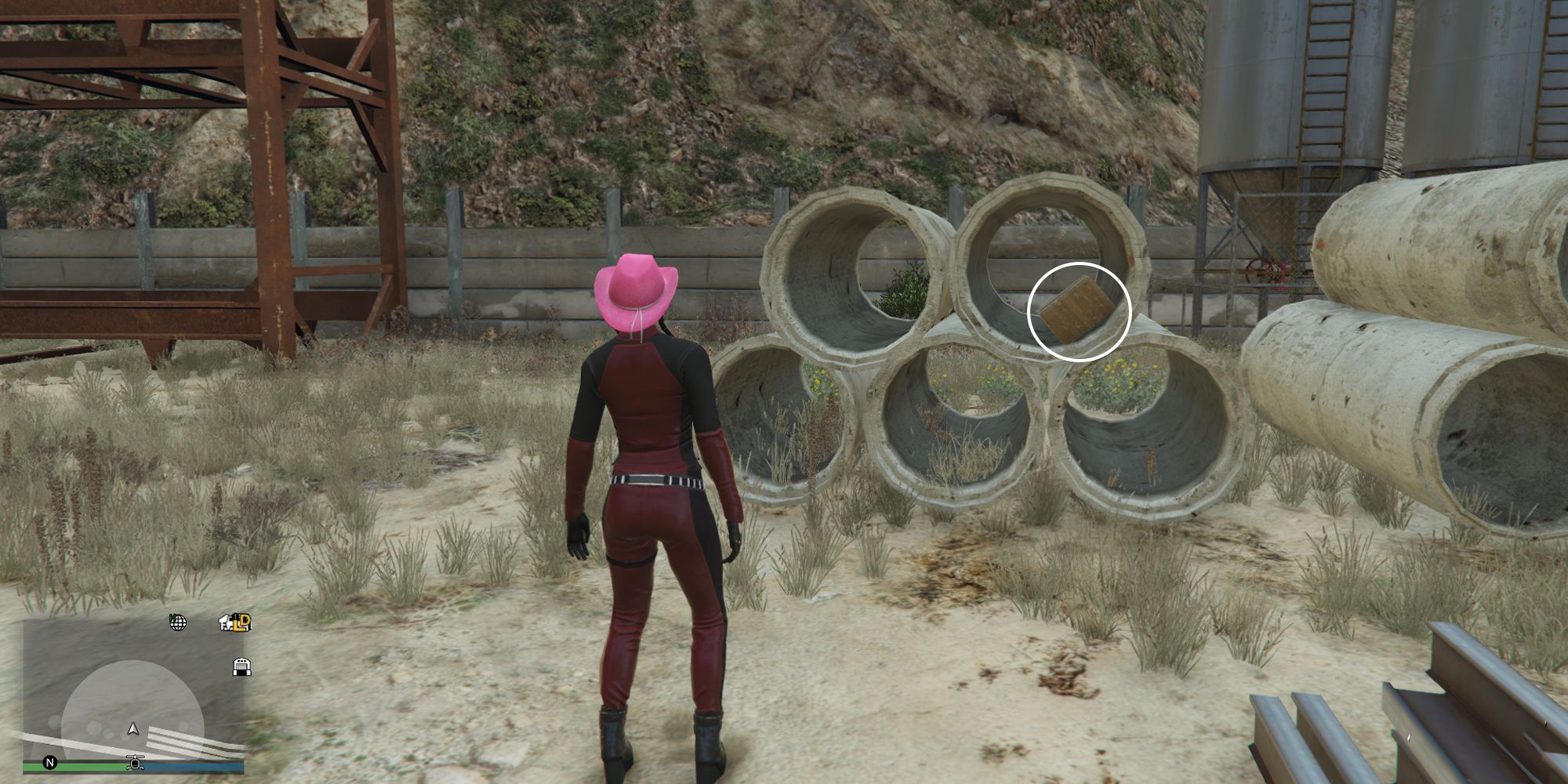 Image depicts a female character in GTA Online at Railyard Clucking Farms stood in front of a stack of concrete pipes. In a pipe on top of the stack on the right-hand side, G's Cache box sits inside.