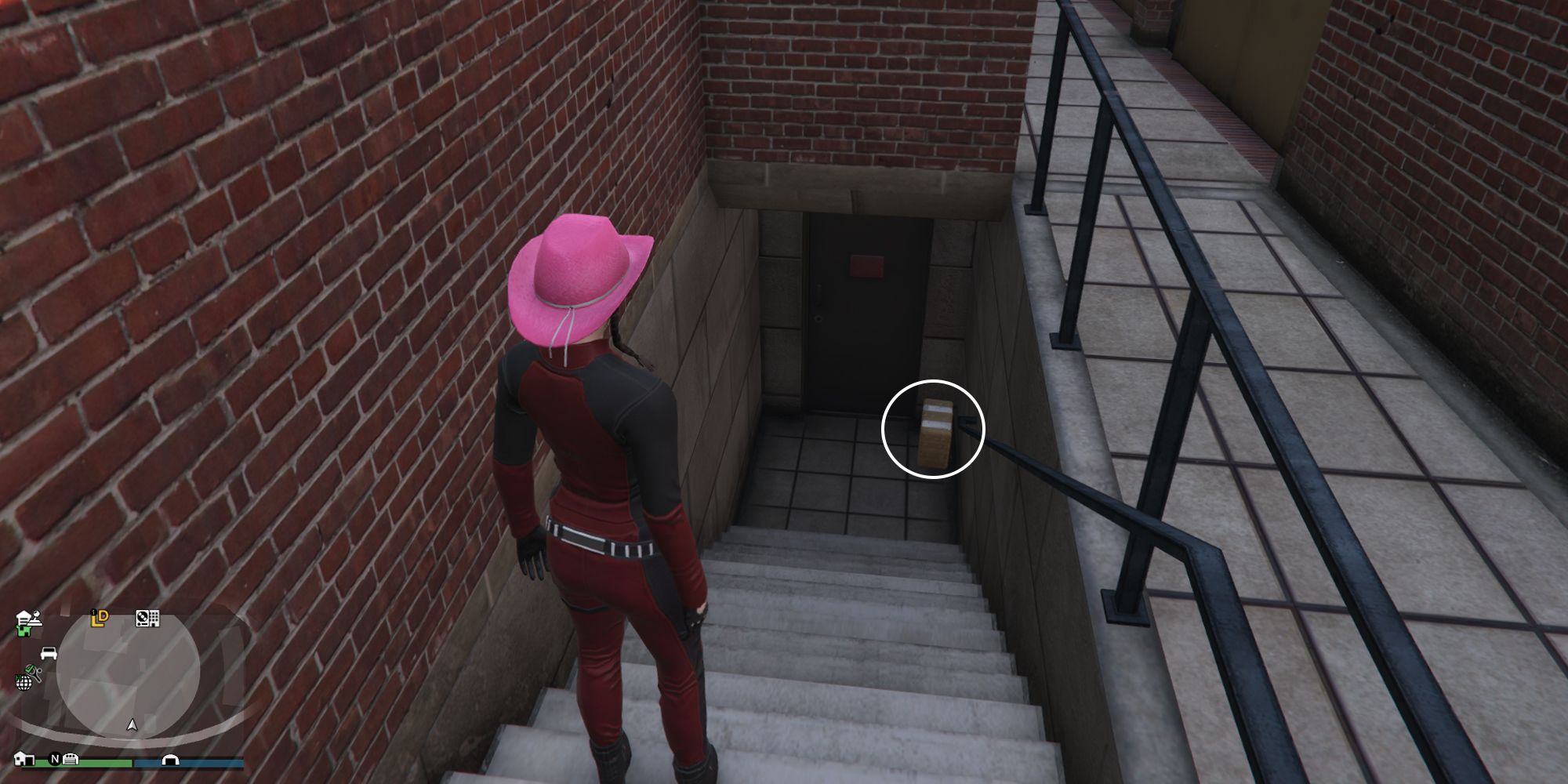 Image depicts a female player in GTA Online at the University of San Andreas stood at the top of a staircase looking down at a door. At the bottom of the staircase, G's Cache is on the floor next to the door.