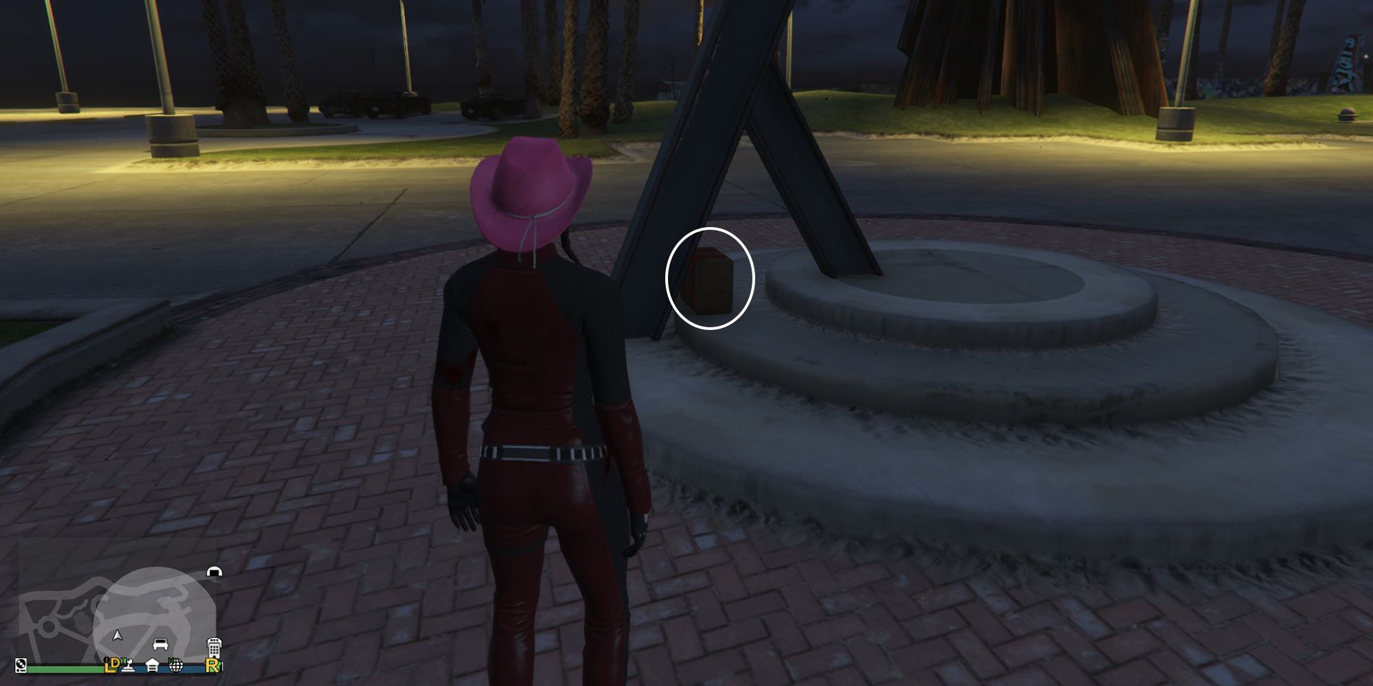 Image depicts a female player in GTA Online at Vespucci Beach Skate Park facing a metal sculpture that splits into a 'Y' shape at the bottom. This is on a circular concrete platform that has three tiers of circles. In the gap between the two metal parts, G's Cache is on the ground.
