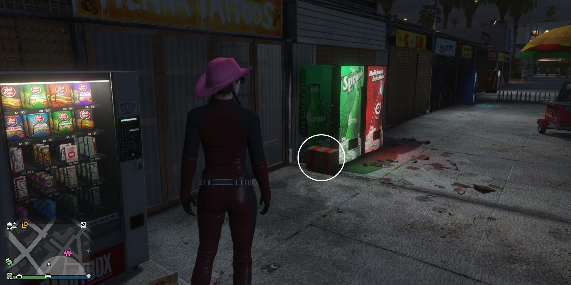 Image depicts a female player in GTA Online at Vespucci Beach Skate Park stood next to the 'Henna Tattoos' Shop on the main strip. There are three vending machines in front of the tattoo shop, and G's Cache is on the floor next to a green one selling drinks.