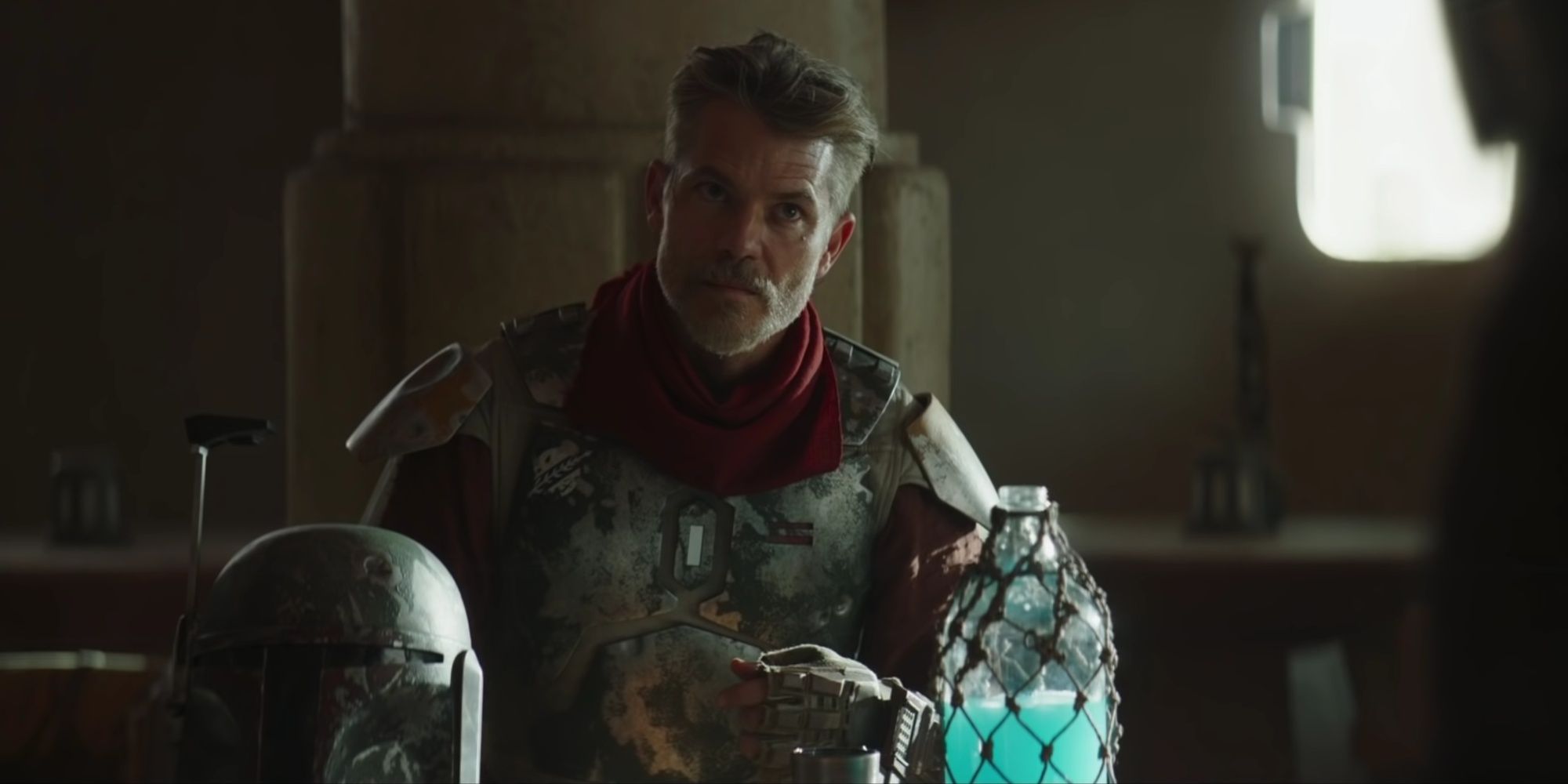 Cobb Vanth sitting at a table with Boba Fett's helmet and a flask of a blue drink on the table being confronted by the Mandalorian.