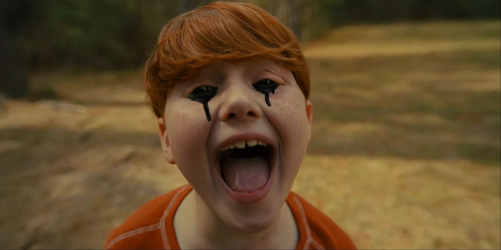 Close-up of a red-haired boy with black tears coming from his eyes mouth open and screaming in a park field.