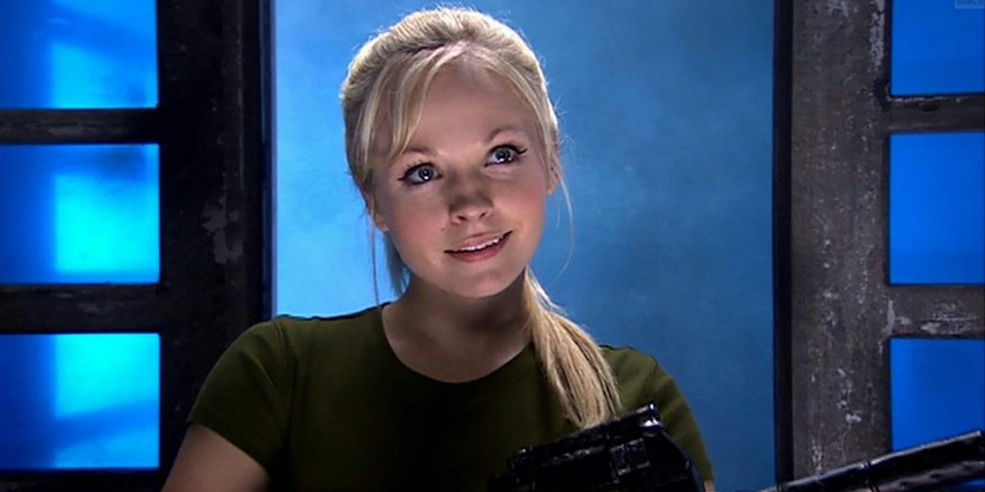 Jenny, the doctor's daughter in Doctor Who stands smiling in front of a blue background
