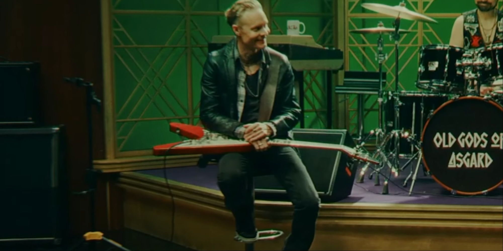 Live-action Bob Baldur sitting by the stage looking at his other band members in the studio of In Between with Mr. Door, hands resting on his red guitar.