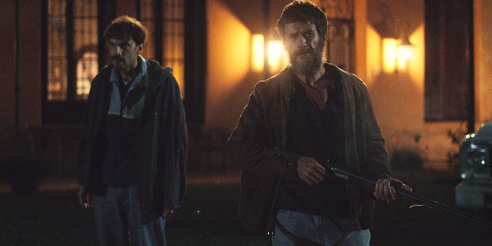 A shot from When Evil Lurks - two men stand outside a lit house at night. The man on the right wields a rifle.