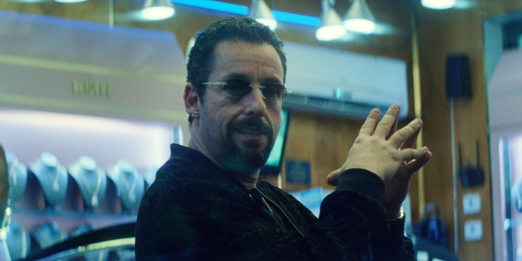Adam Sandler's Howard Ratner with his hands clasped together staring toward the camera inside his jewelry store, with a devious look.