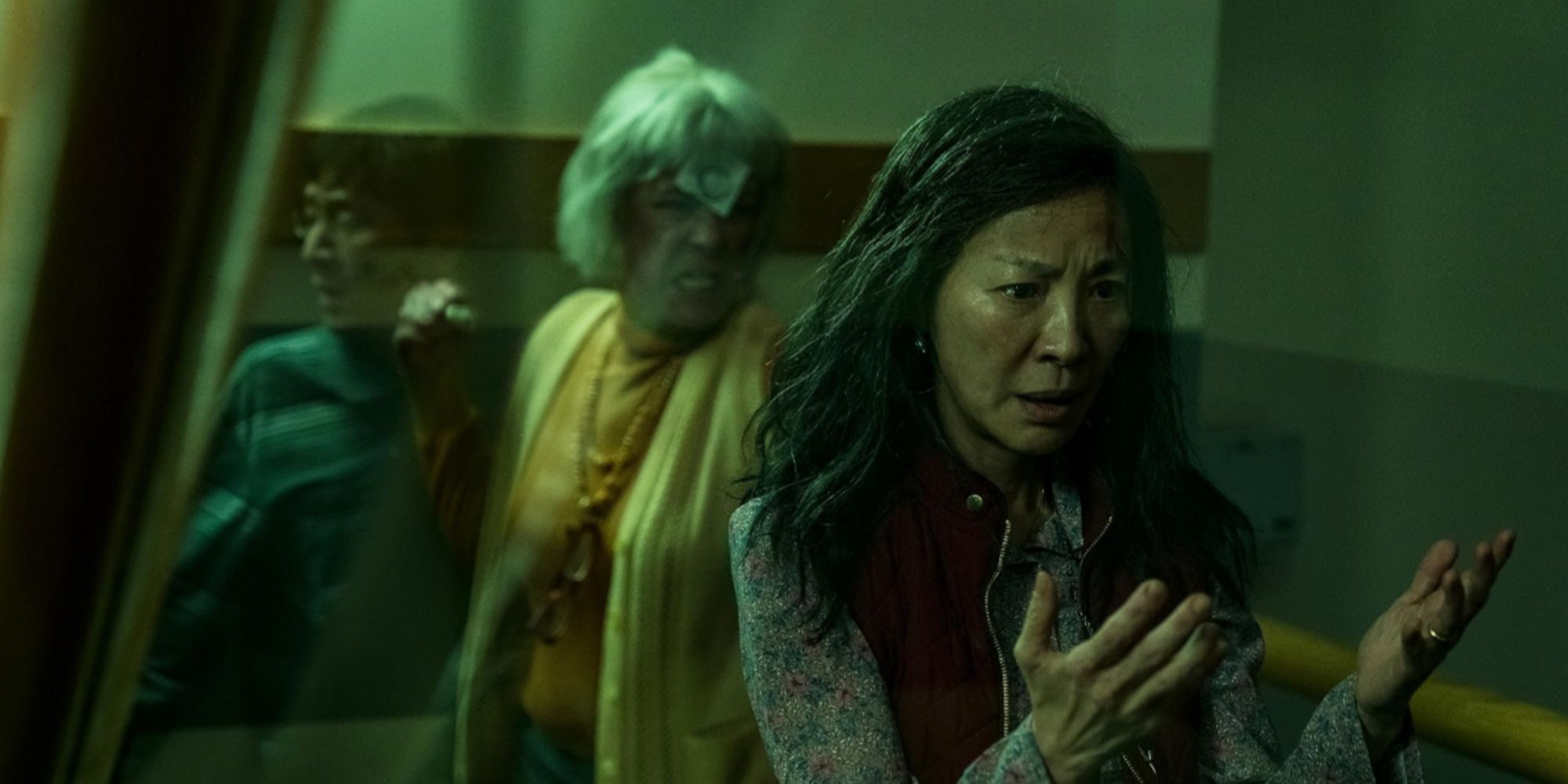 Evelyn looking at her hands with an angry Alpha Dierdre behind her and Waymond in the reflection of the glass.