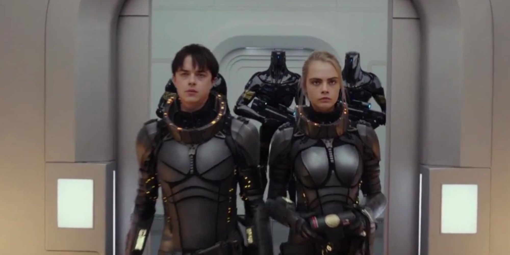 Valerian and his partner Laureline walking side-by-side in their black space suits, escorted by armed robots.