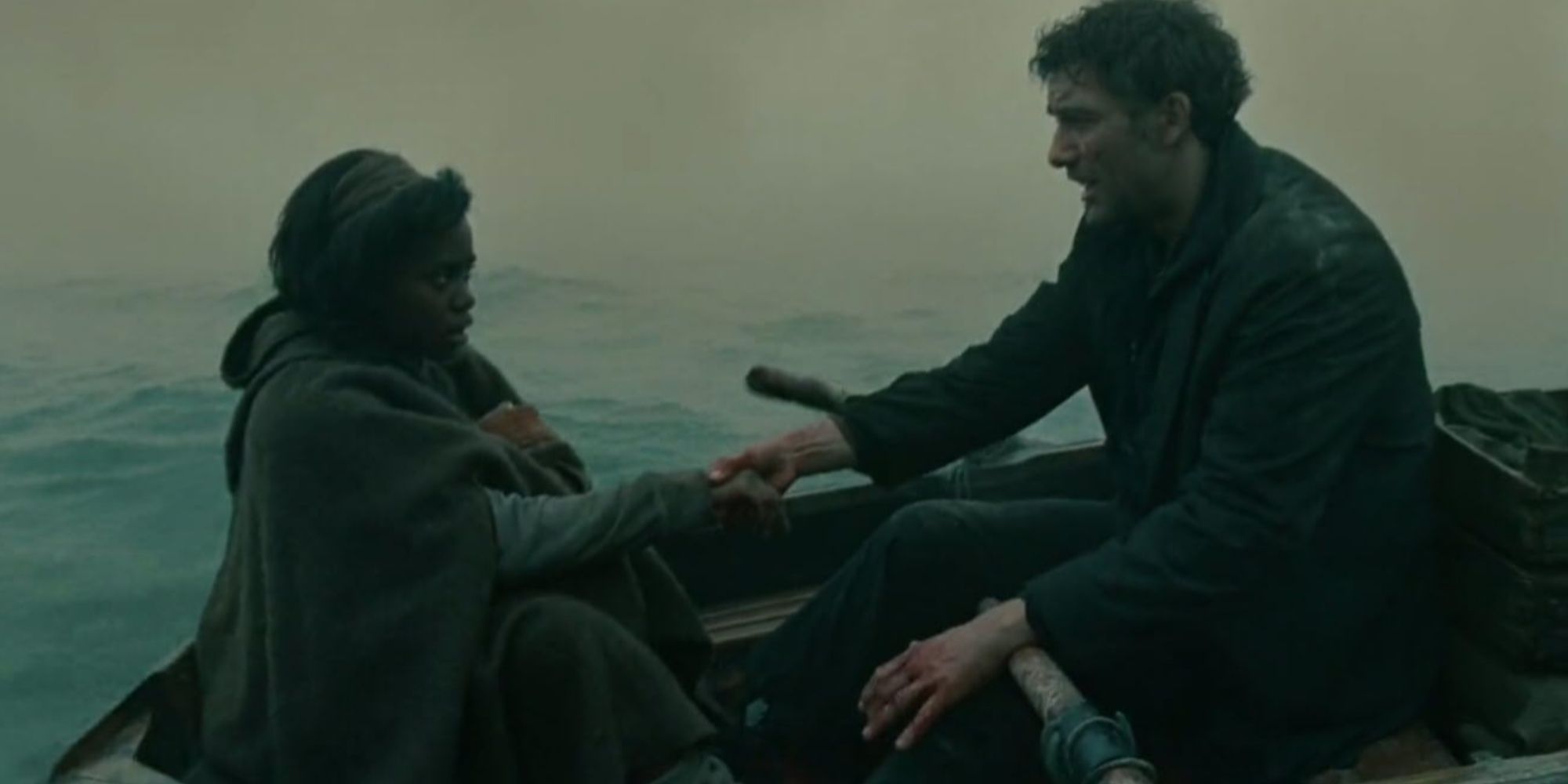 Kee wrapped in a coat holding onto Theo Faron's hand as they row away on a boat.
