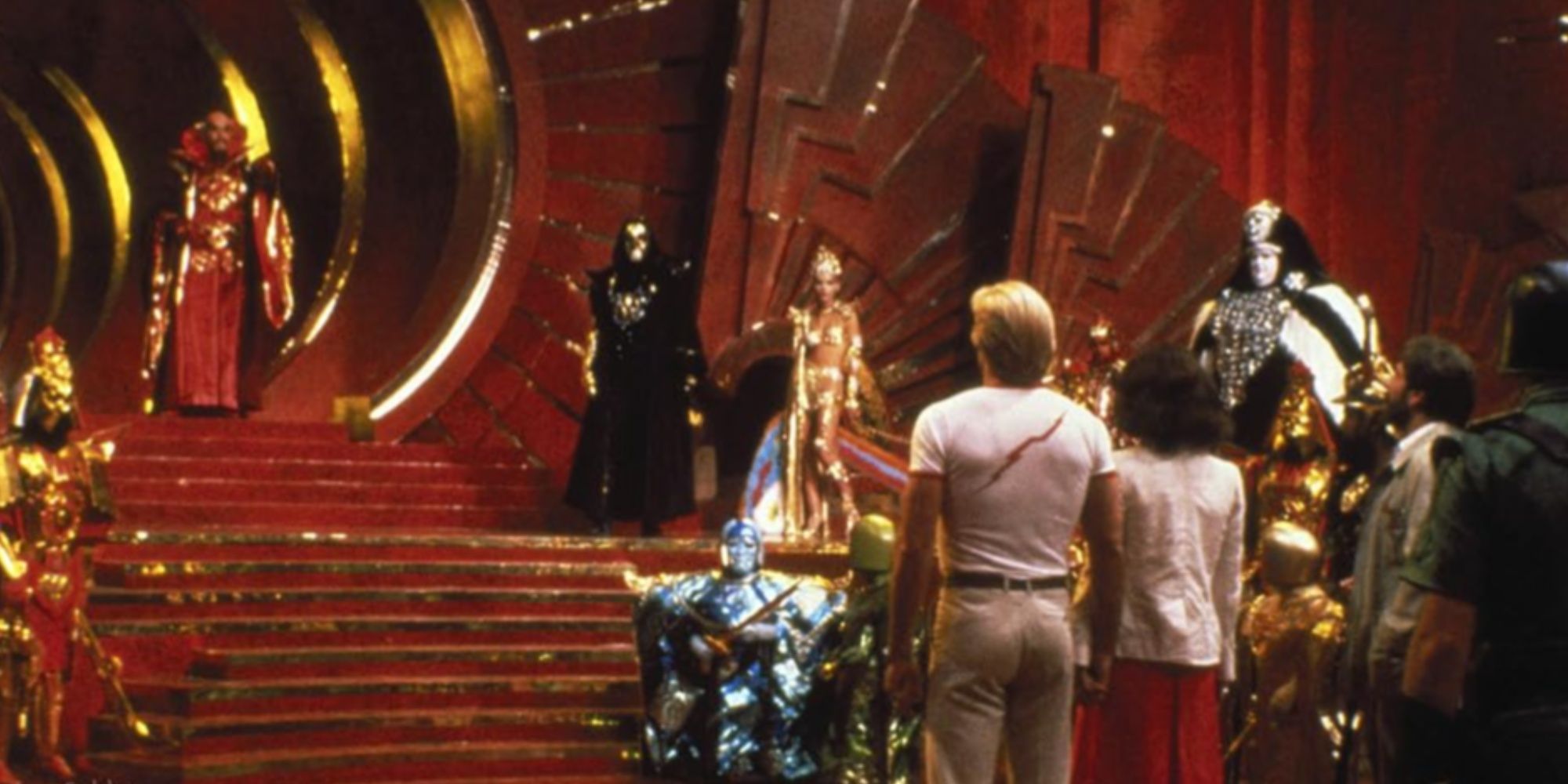 Sam J. Jones' character Flash Gordon standing beside actress Melody Anderson confronting Emperor Ming portrayed by Max von Sydow, who's walking down stairs in a red-yellow room.