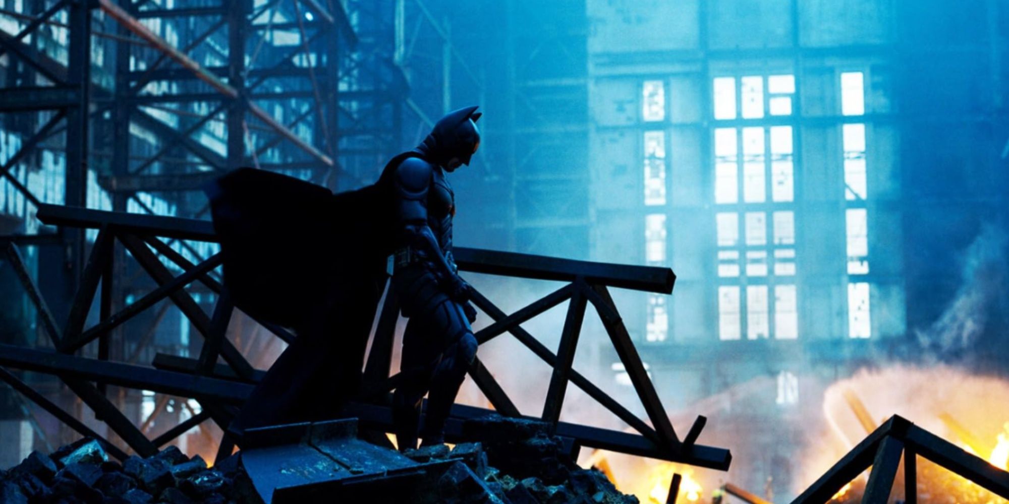 Christian Bale's Batman standing on rubble with his head down in a shot that features blues and orange from the fire in the distance.
