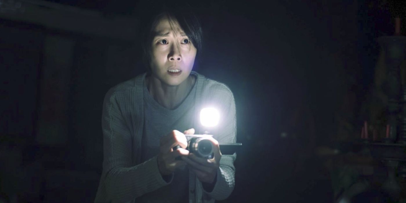 The protagonist of Incantation holds a camera and flashlight, she looks scared.
