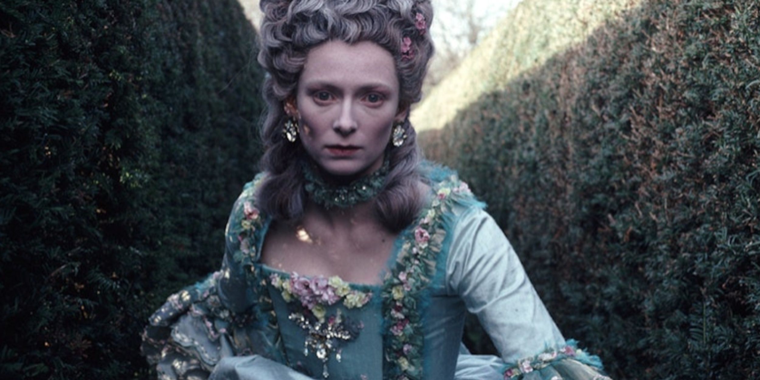 Tilda Swinton moves through a hedge maze in the movie Orlando, dressed in a ballgown and wearing an elaborate wig.