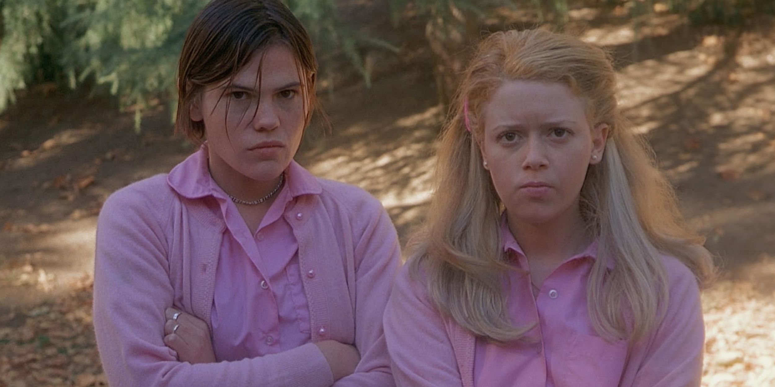 Natasha Lyonne and Clea DuVall in But I'm A Cheerleader, standing in the woods wearing entirely pink outfits. Both women look unhappy.