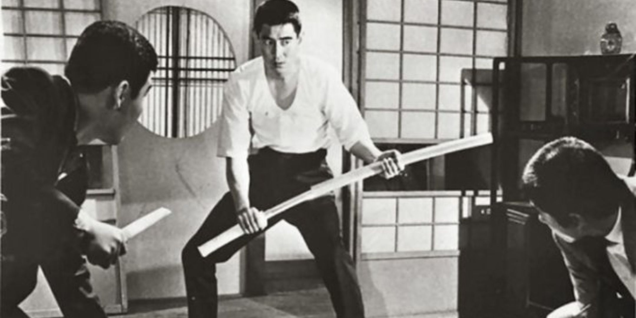 Ken in a movie holding a sword while two attackers wait for him to make a move.
