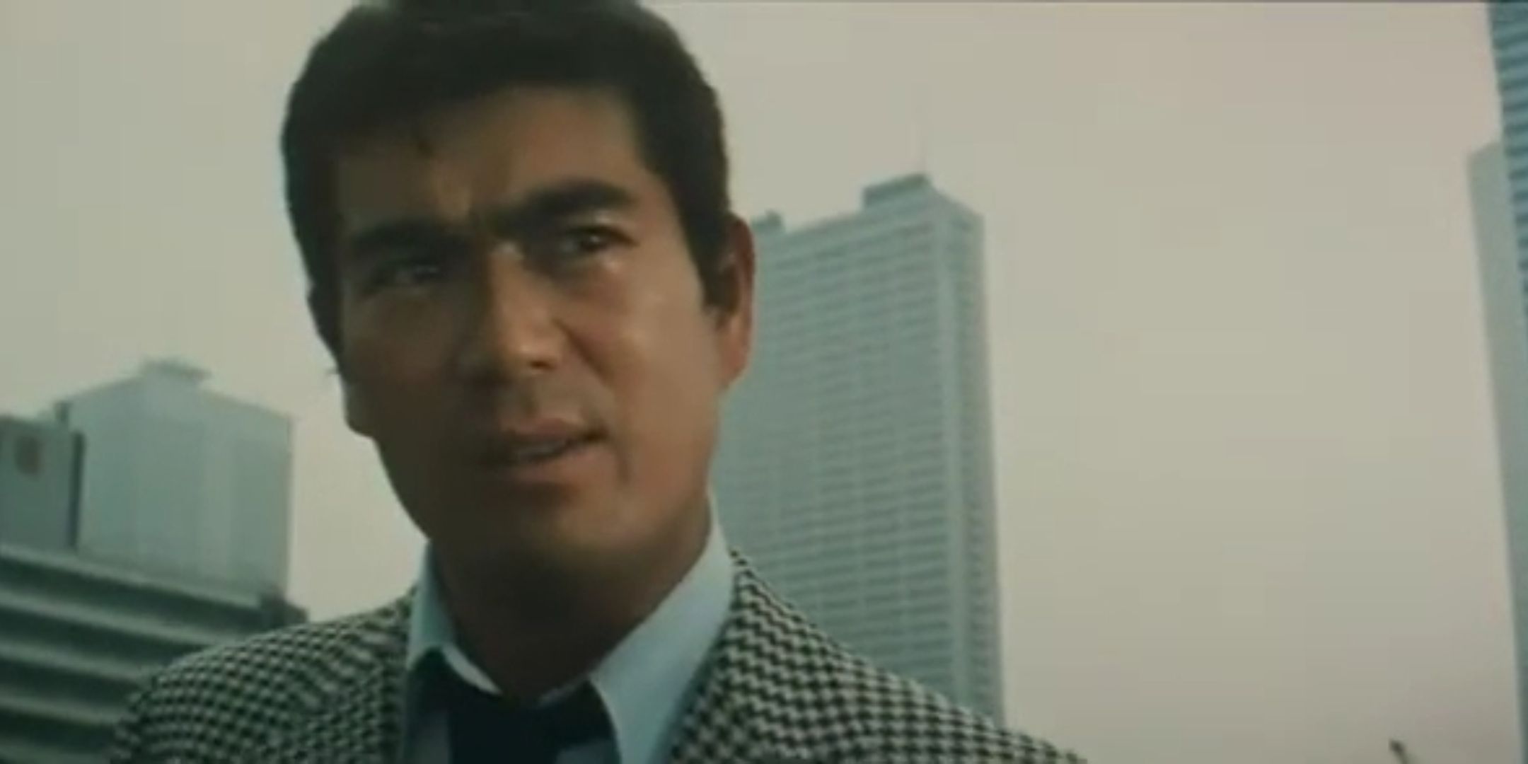 A closeup of Ken Takakura in a crime drama, he is wearing a plaid suit and there is a skyscraper in the distance.