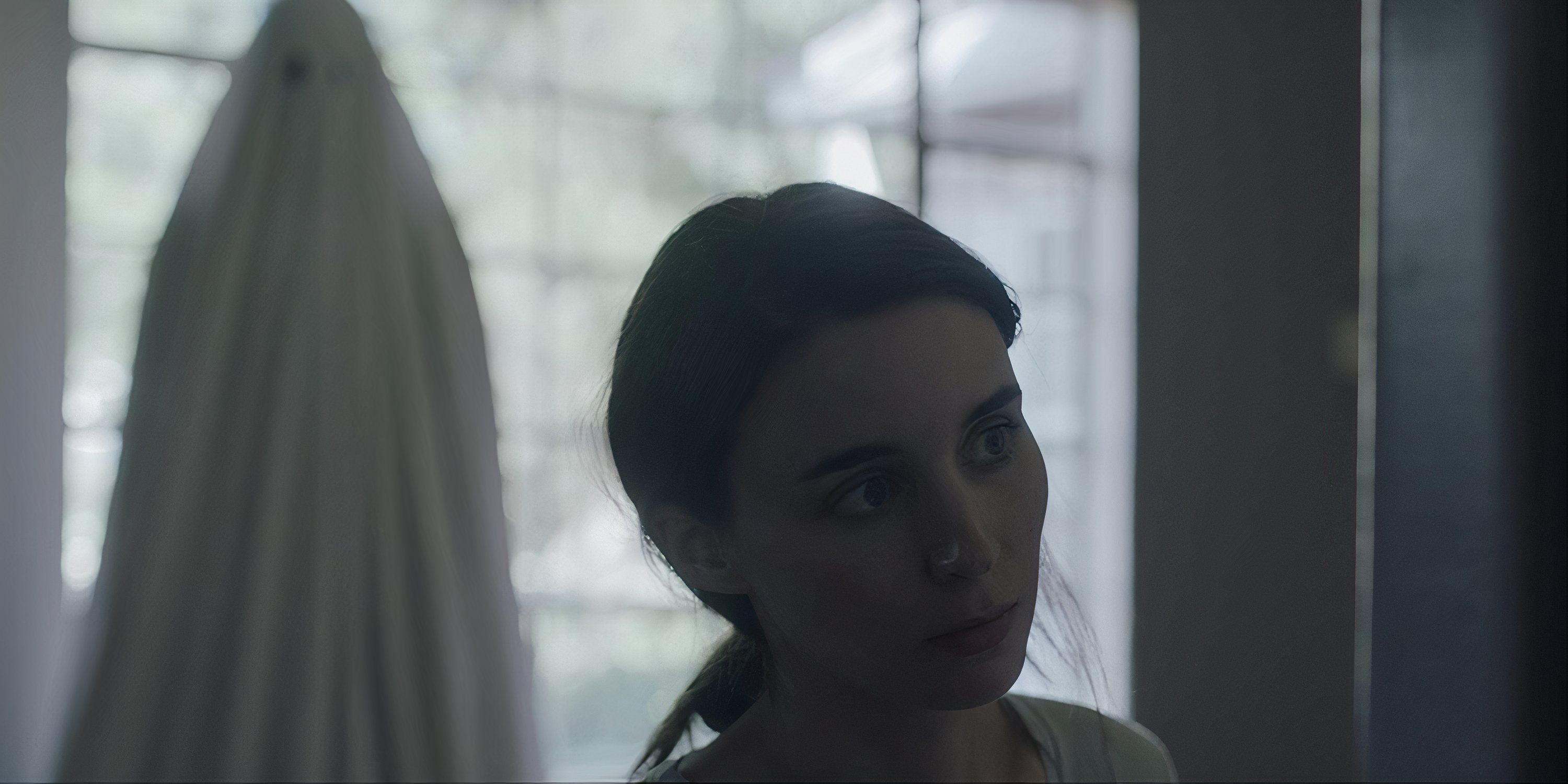 Rooney Mara looking ahead as a ghost in a white sheet watches her from behind in A Ghost Story.