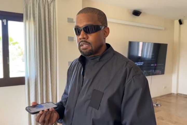 Kanye West standing in a mostly empty room, speaking to someone on the phone while wearing sunglasses