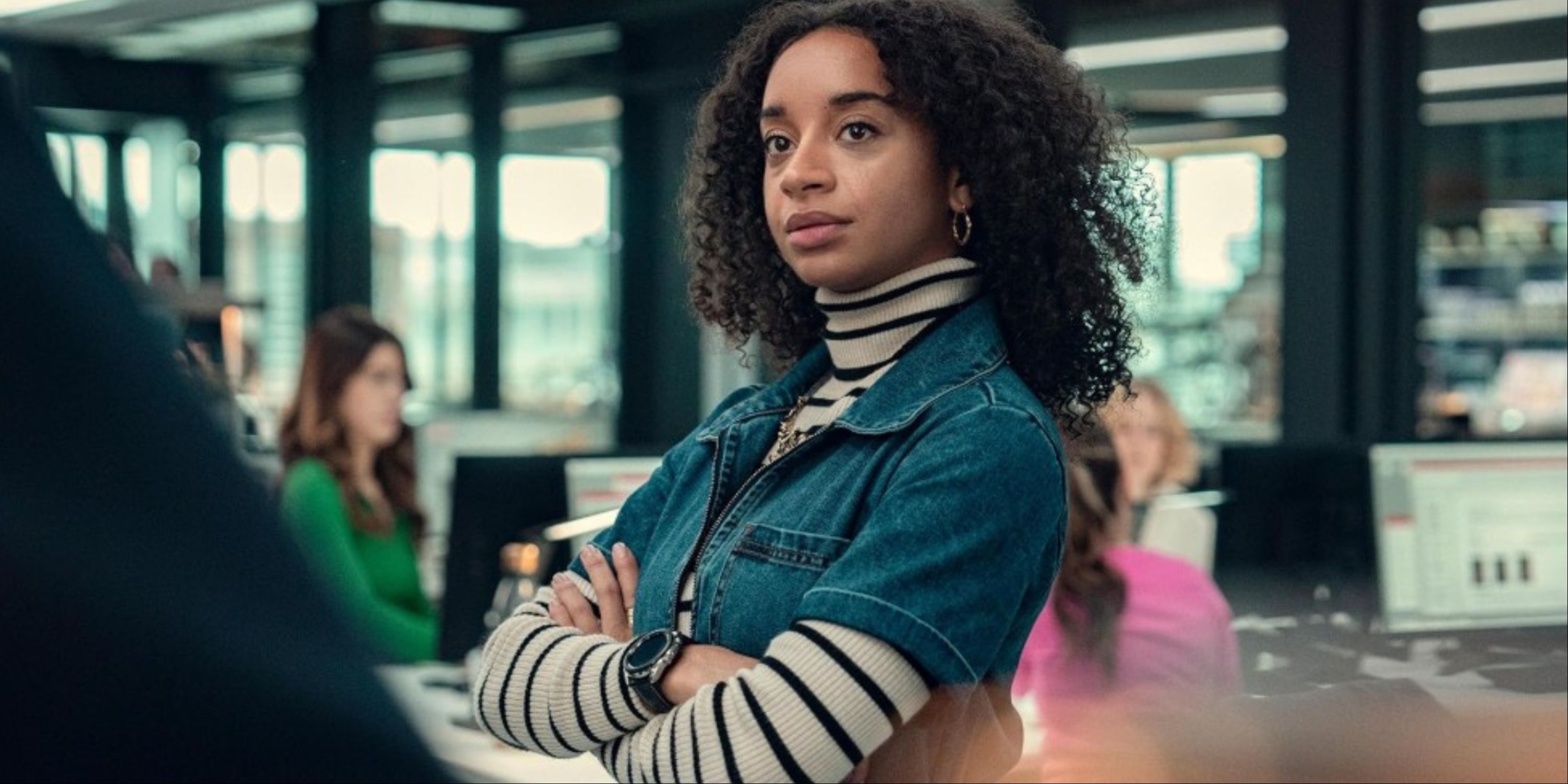 Siena Kelly crossing her arms as her character Maria, with Verity's faces faded in the background at her desk in Black Mirror's Bete Noire episode.