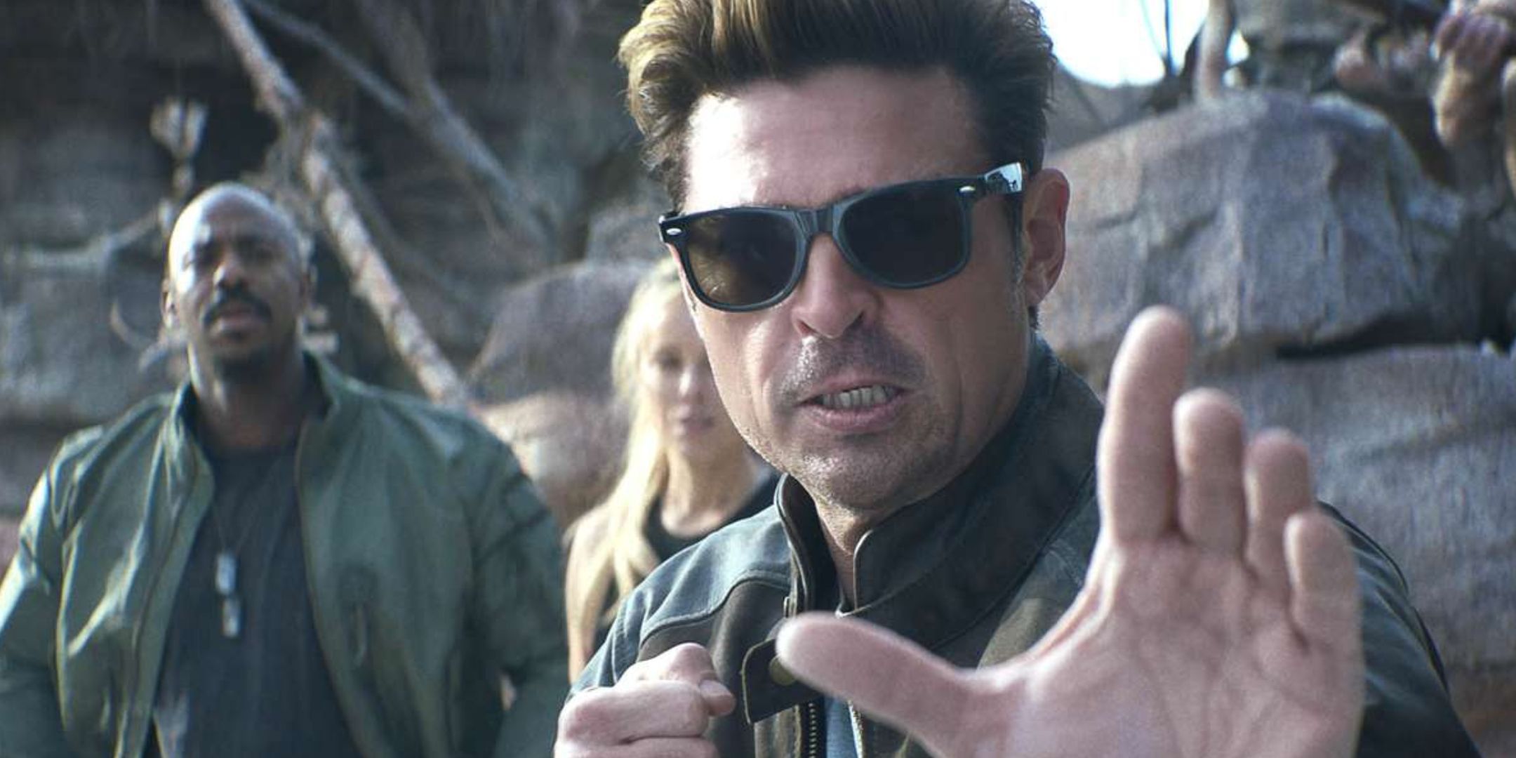A close up of Johnny Cage, as played by Karl Urban in Mortal Kombat 2. He is wearing sunglasses and pulling a martial arts pose. Two people are stood behind him, and rubble can be seen in the background.