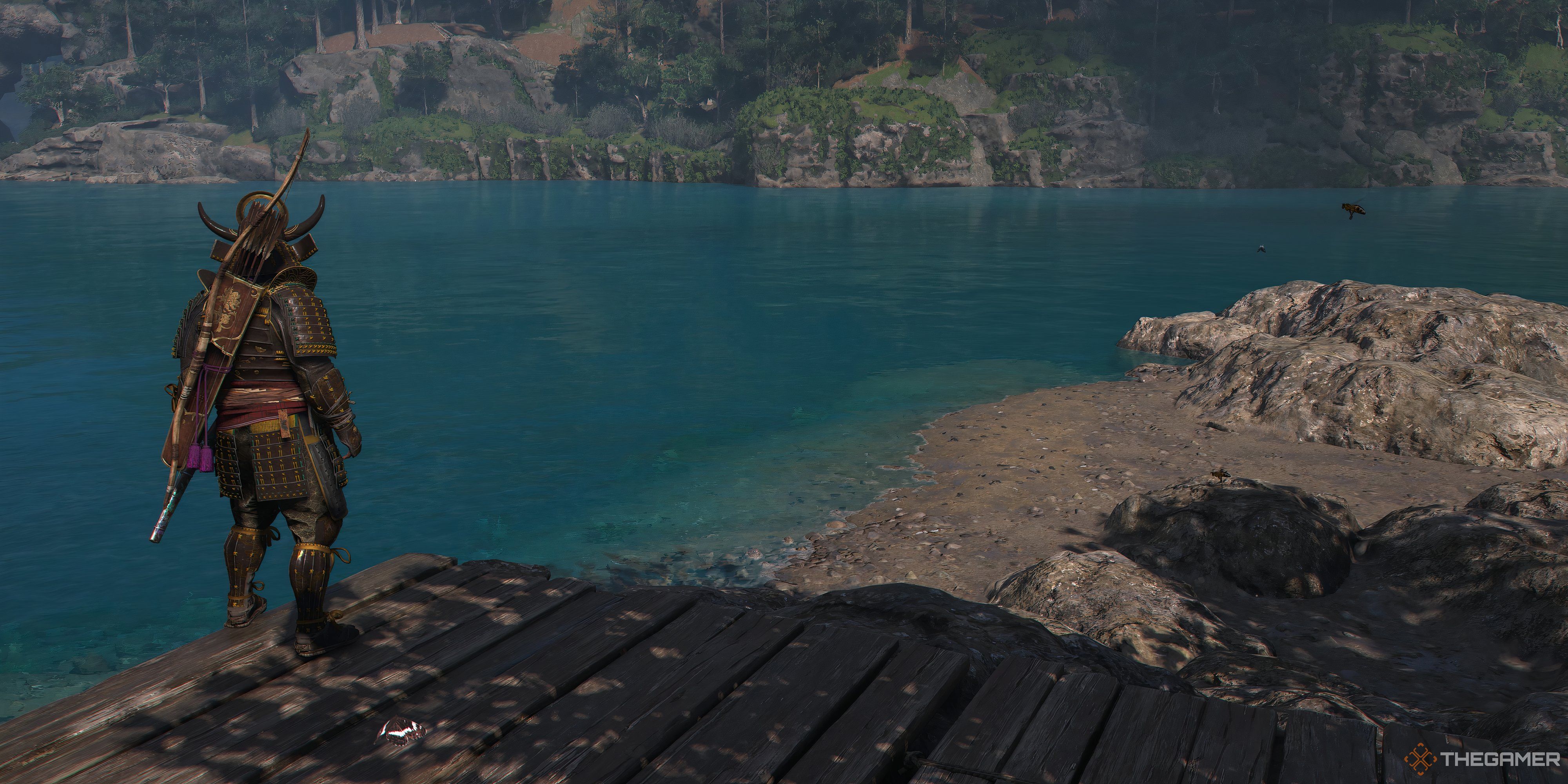 Asssassins Creed Shadows Yaskue facing the water with trees and rocks on the other side. He is on a wooden platform.