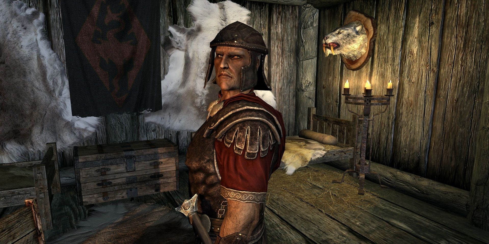 A man dressed in red and metal armor stares at the viewer inside his bunk with a banner and animal head on the walls.
