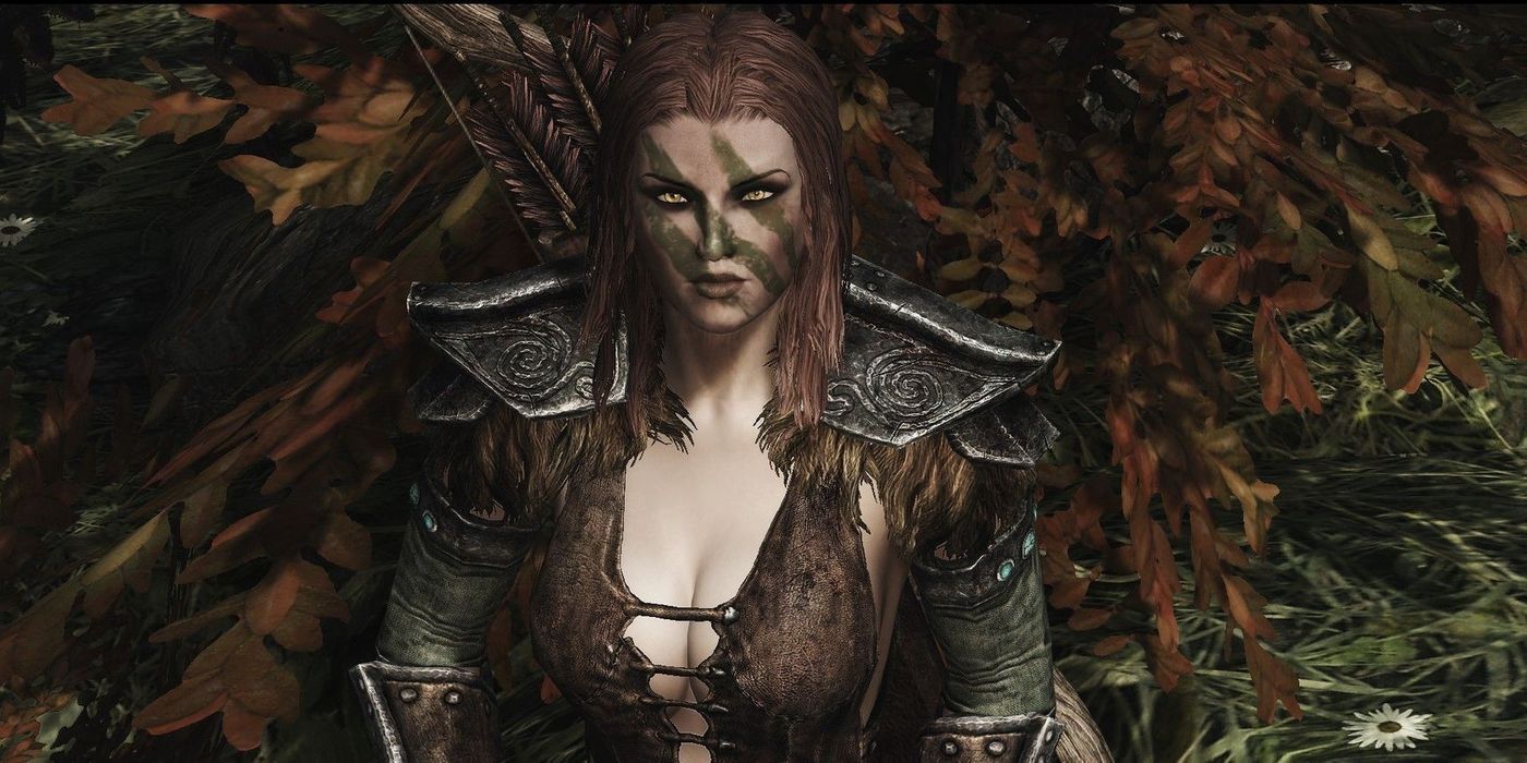A woman dressed in ancient armor and green face paint stares at the camera with green foliage in the background.