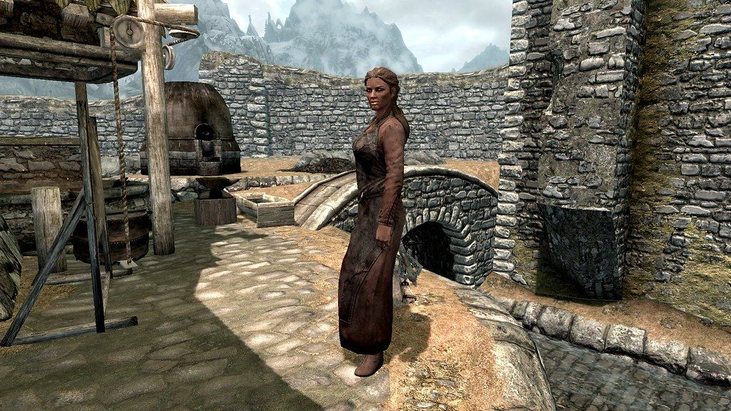 A female blacksmith stands by her forge with a city's walls in the background.